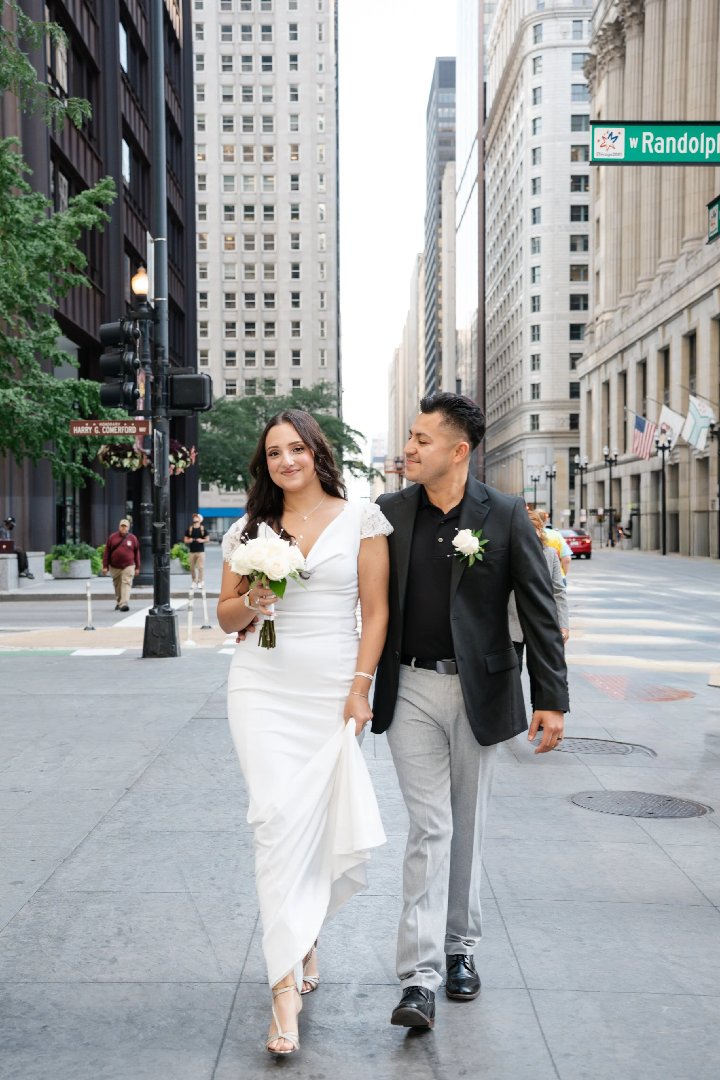 A bride and groom walking in an urban city street, holding bouquets of white flowers, with tall buildings and city street signs behind them.