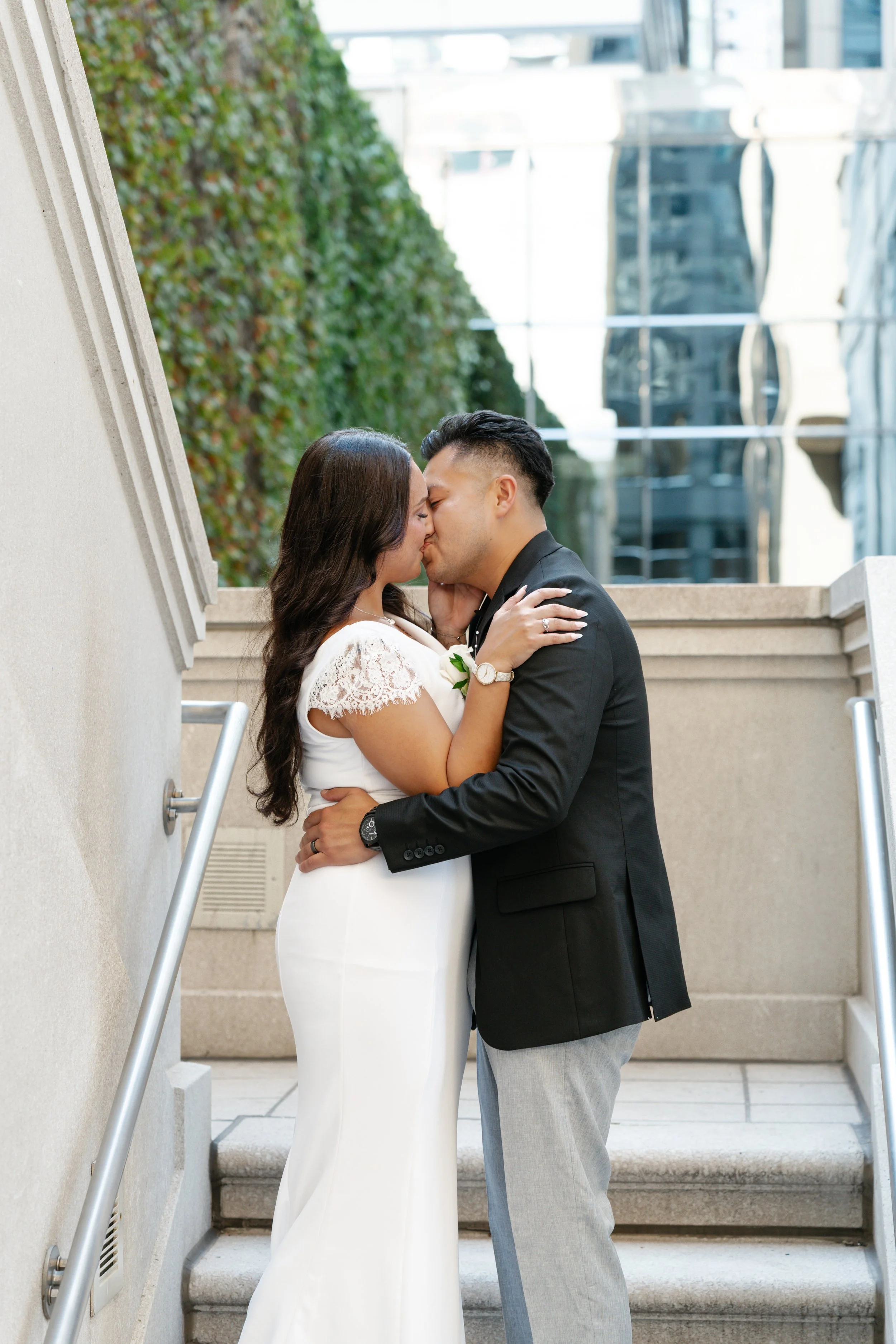 A couple kissing on staircase outdoors, woman in white dress with lace sleeves, man in black blazer and gray pants, city buildings in background.