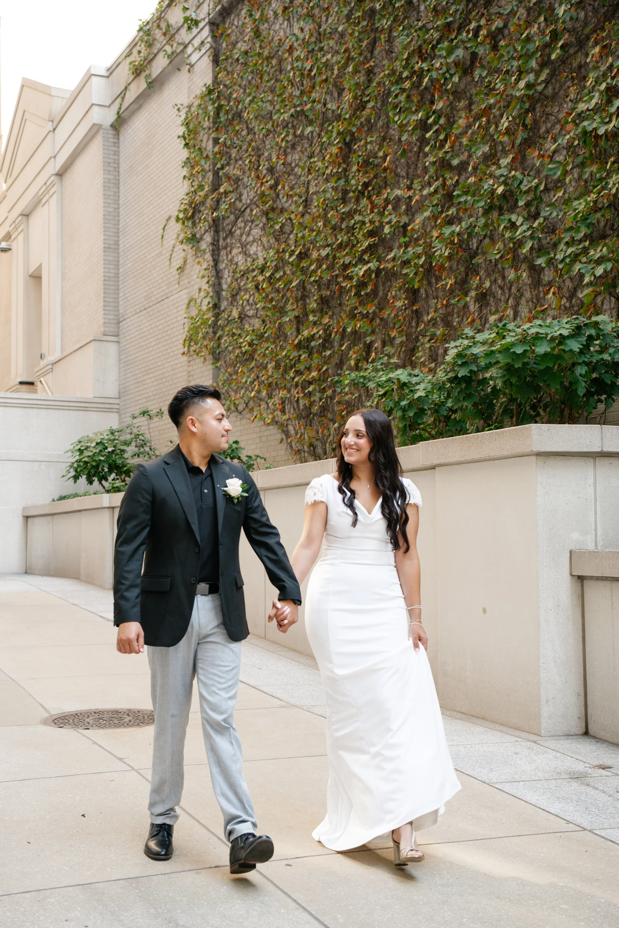 A newlywed couple walking hand in hand outdoors, smiling at each other. The man is in a black blazer, black shirt, and light gray pants, with a white boutonniere. The woman is in a white wedding gown, with long dark hair and high heels.
