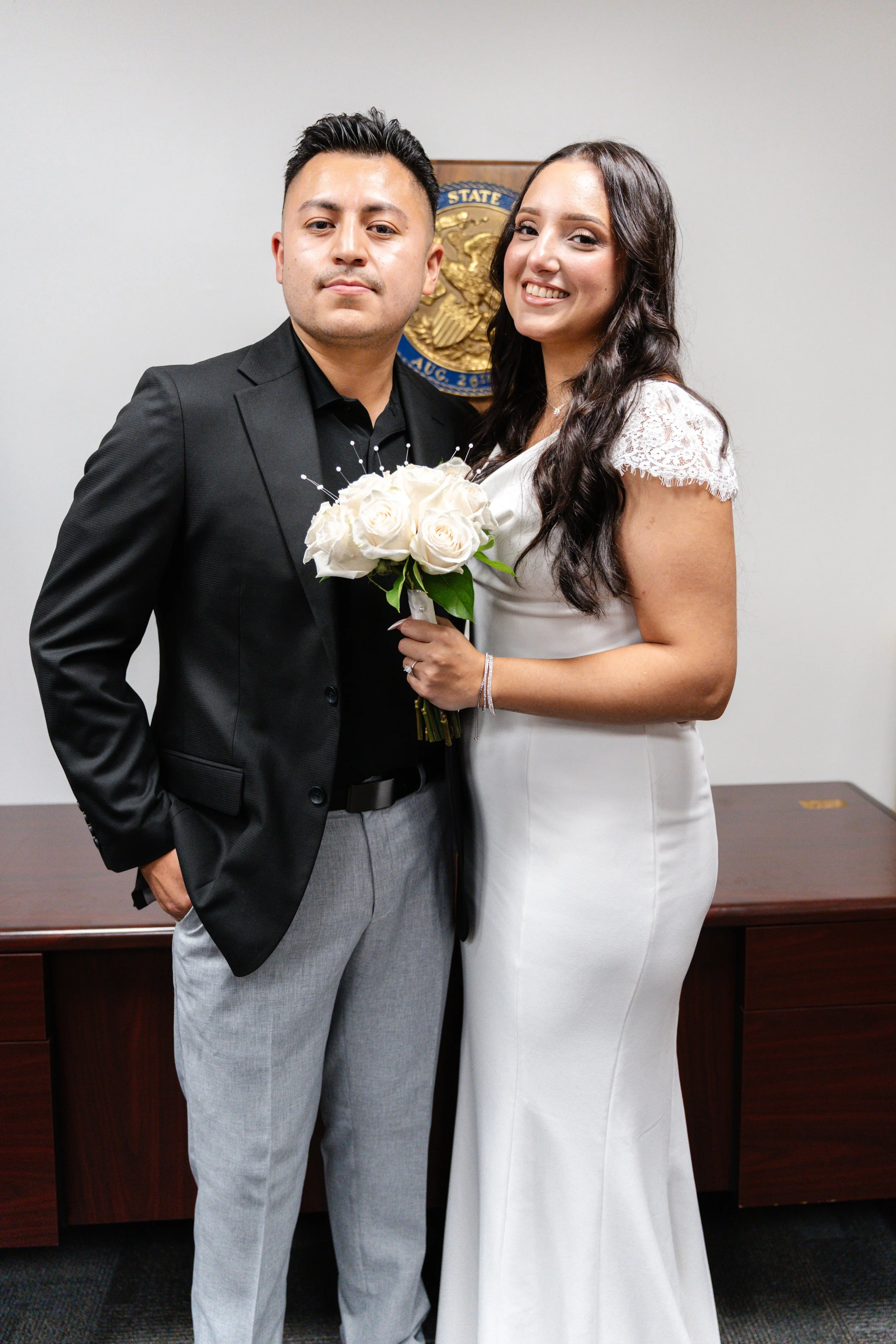 A couple in formal attire standing indoors. The woman, in a white wedding dress, holds a bouquet of white roses. The man, in a black blazer and gray pants, stands beside her. Behind them is a wall with a crest or emblem.