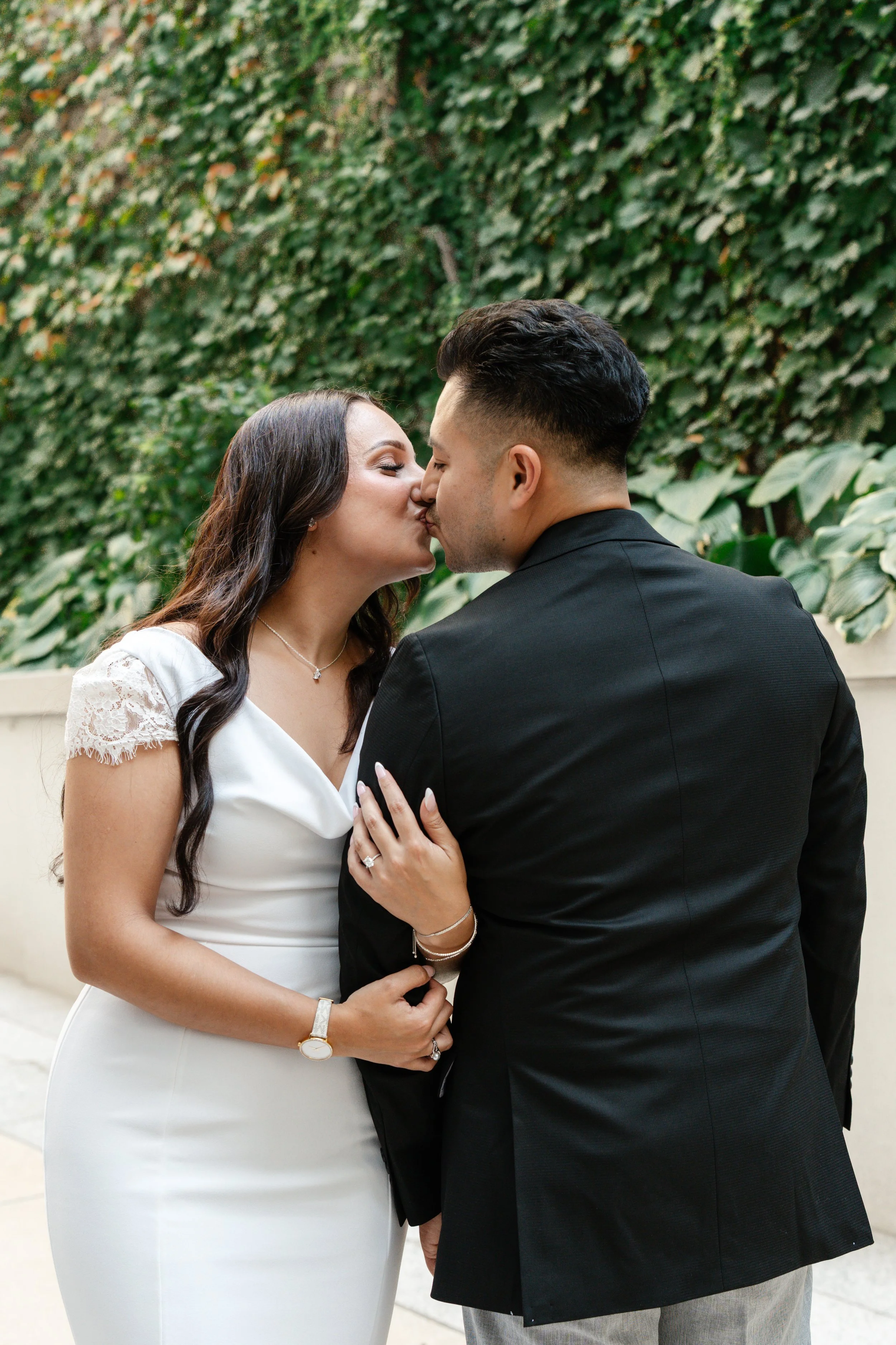 A couple dressed in wedding attire sharing a kiss outdoors, with a green leafy background.
