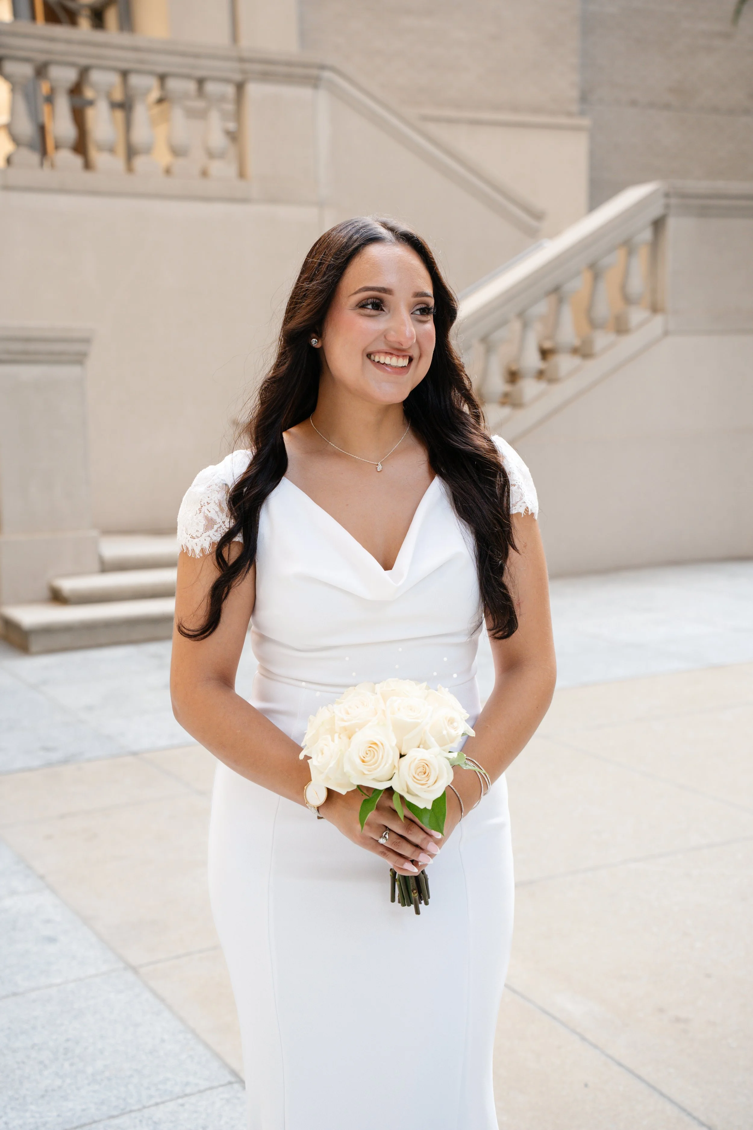 A smiling bride in a white wedding dress holding a bouquet of white roses, standing in a building with stone stairs and railings.