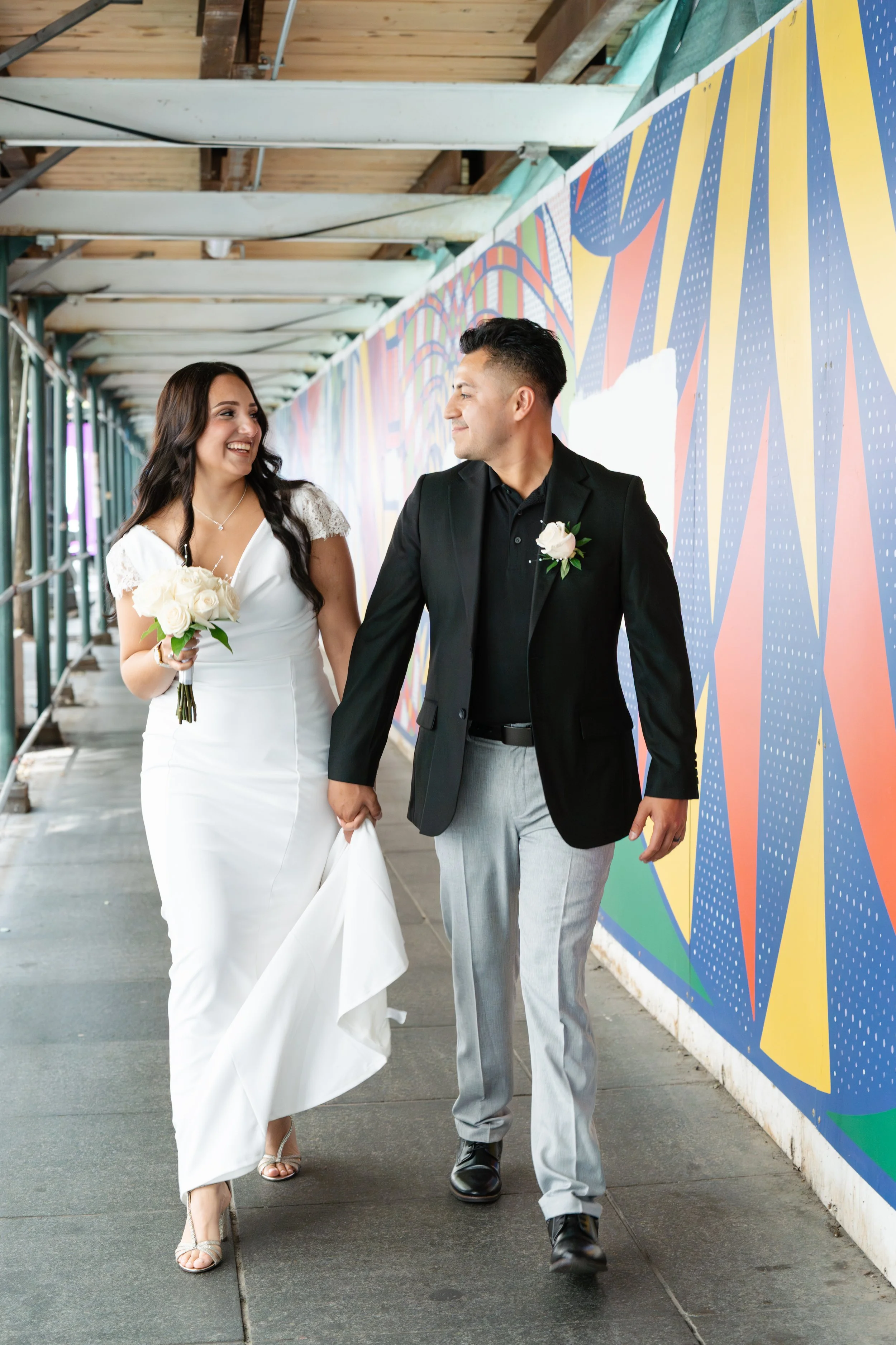 A newlywed couple walking hand in hand under a colorful city bridge, smiling at each other. The bride is holding a bouquet of white roses, wearing a white wedding dress with lace details, and the groom is dressed in a black blazer, black shirt, and l