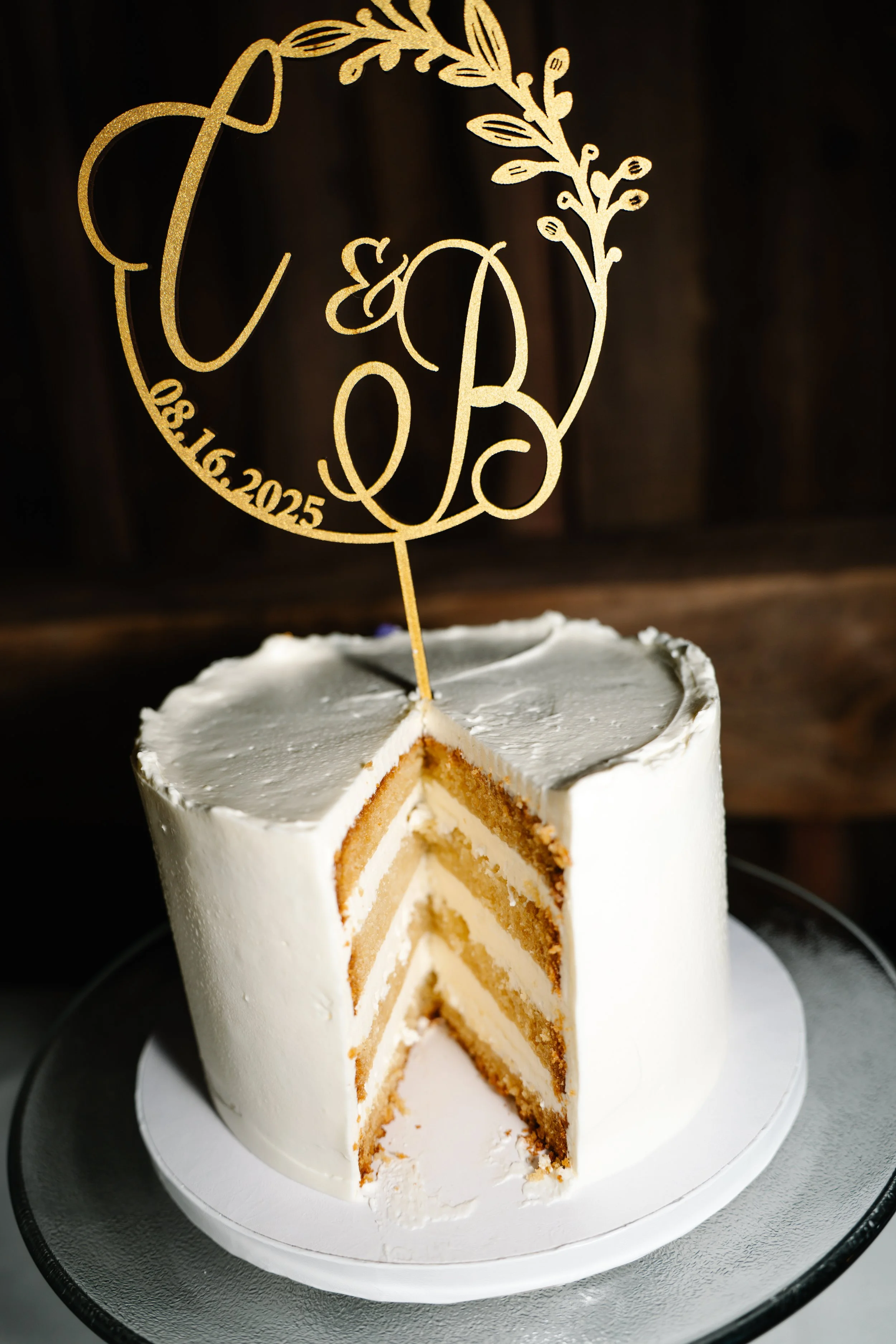 A white wedding cake with a gold topper that includes the initials C & B, a date 08/16/2025, and decorative leaves. The cake has three layers of sponge and cream filling, with a slice removed showing the interior.