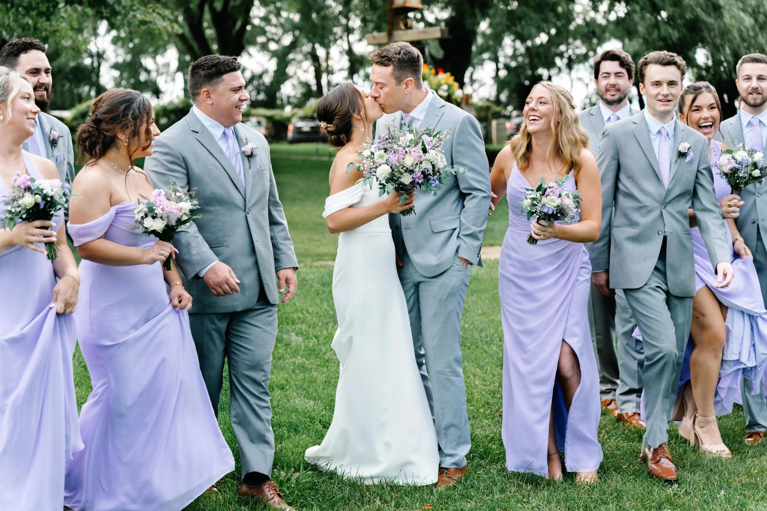 A wedding party outdoors, with the bride and groom kissing, surrounded by bridesmaids and groomsmen holding bouquets, all dressed in formal attire on a grassy lawn.