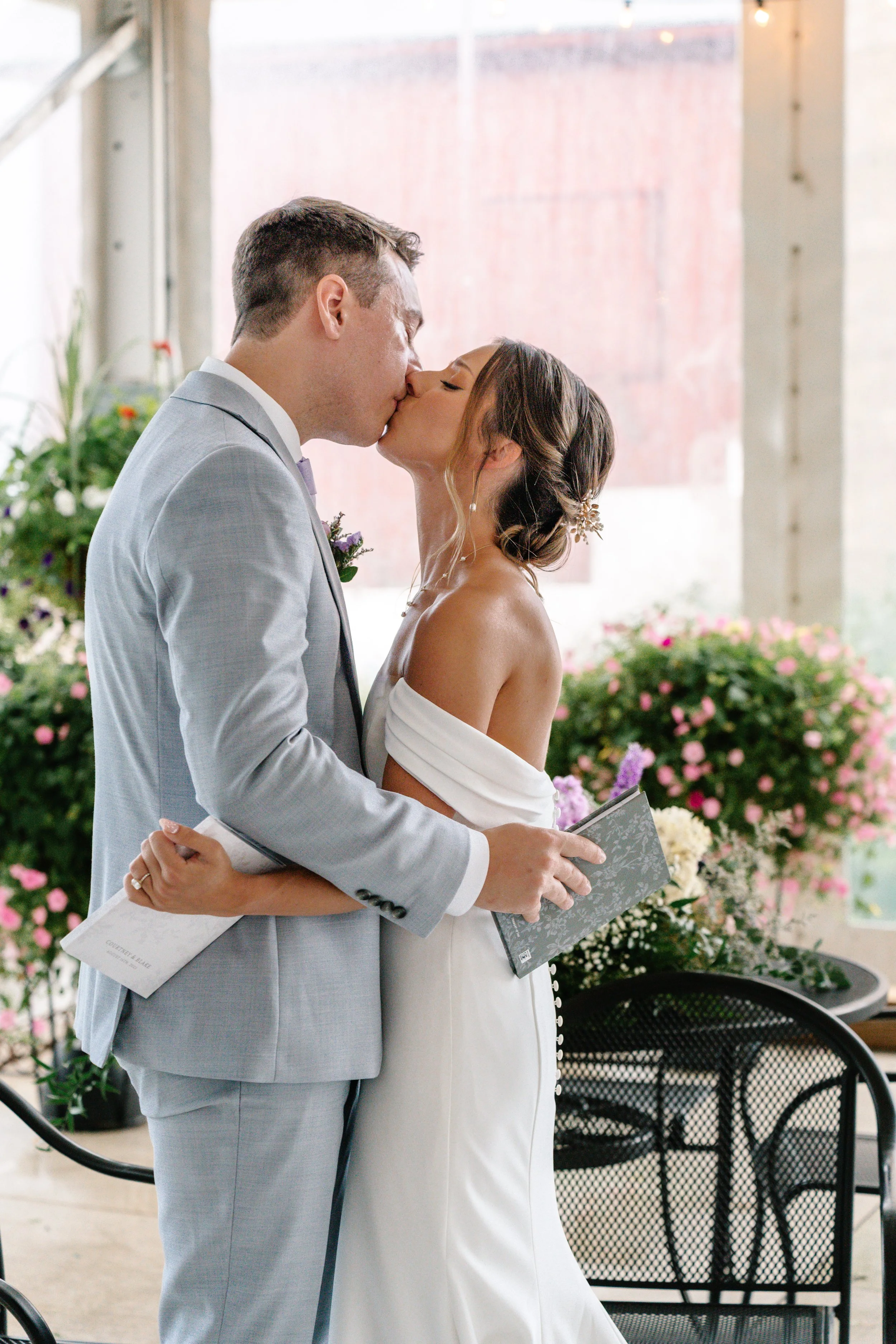 A bride and groom share a kiss during their wedding ceremony, with the bride holding a book and the groom holding a folded paper, surrounded by flowers.