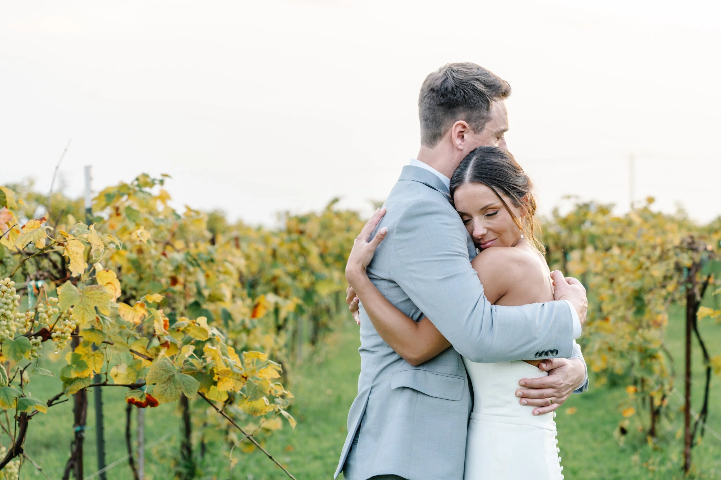 A newlywed couple embracing in a vineyard with green and yellow grapevine leaves in the background.