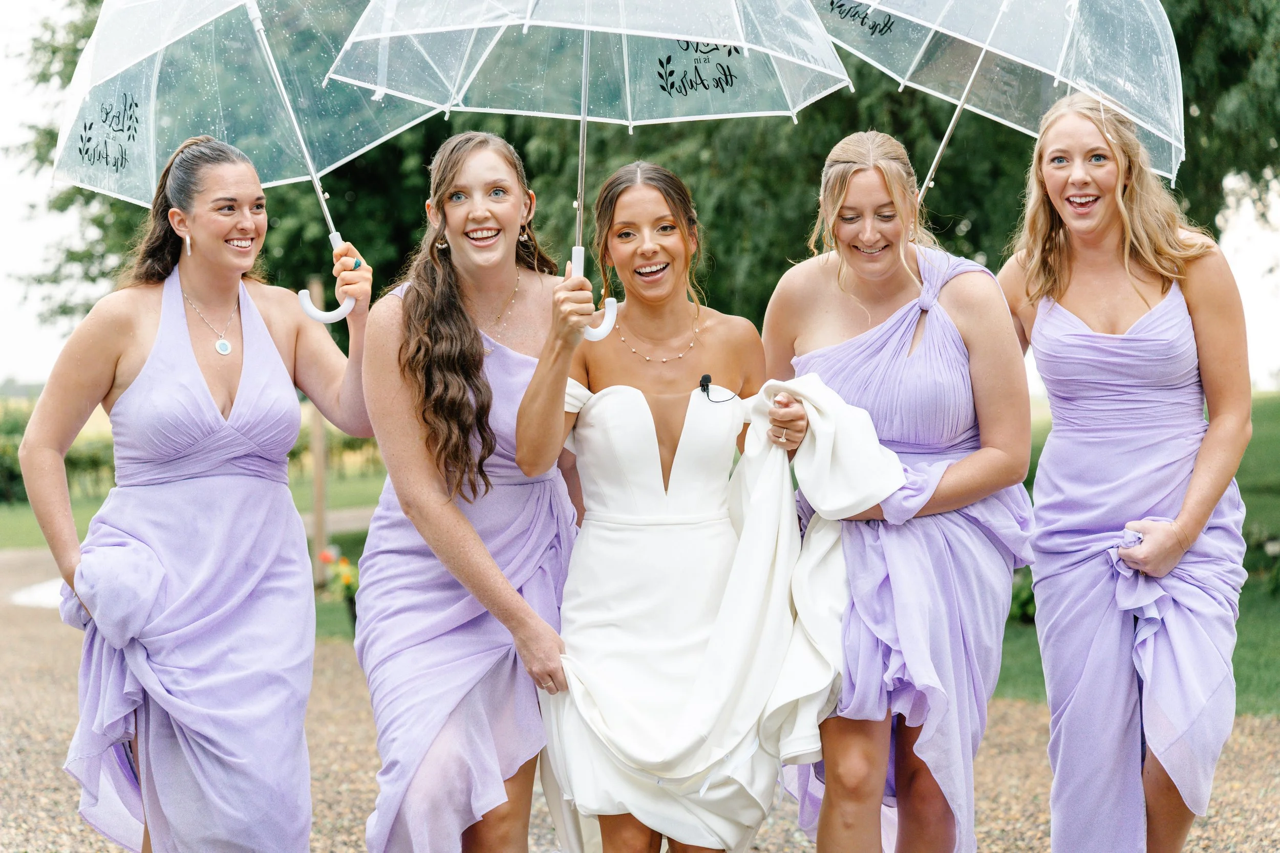 Bride and bridesmaids walking outdoors in the rain, holding umbrellas and smiling, with the bride lifting her dress slightly.