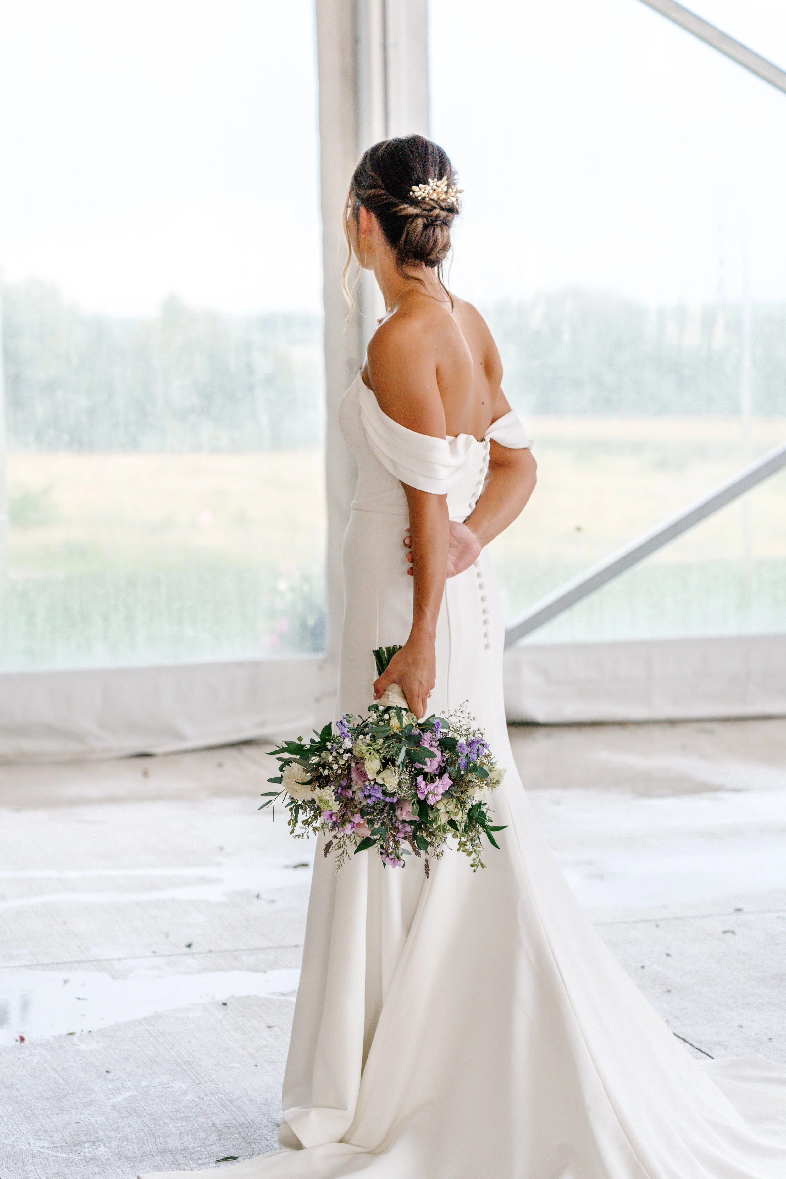 Bride in a white off-shoulder wedding dress holding a bouquet of flowers, standing in front of a window with a view of outdoors.