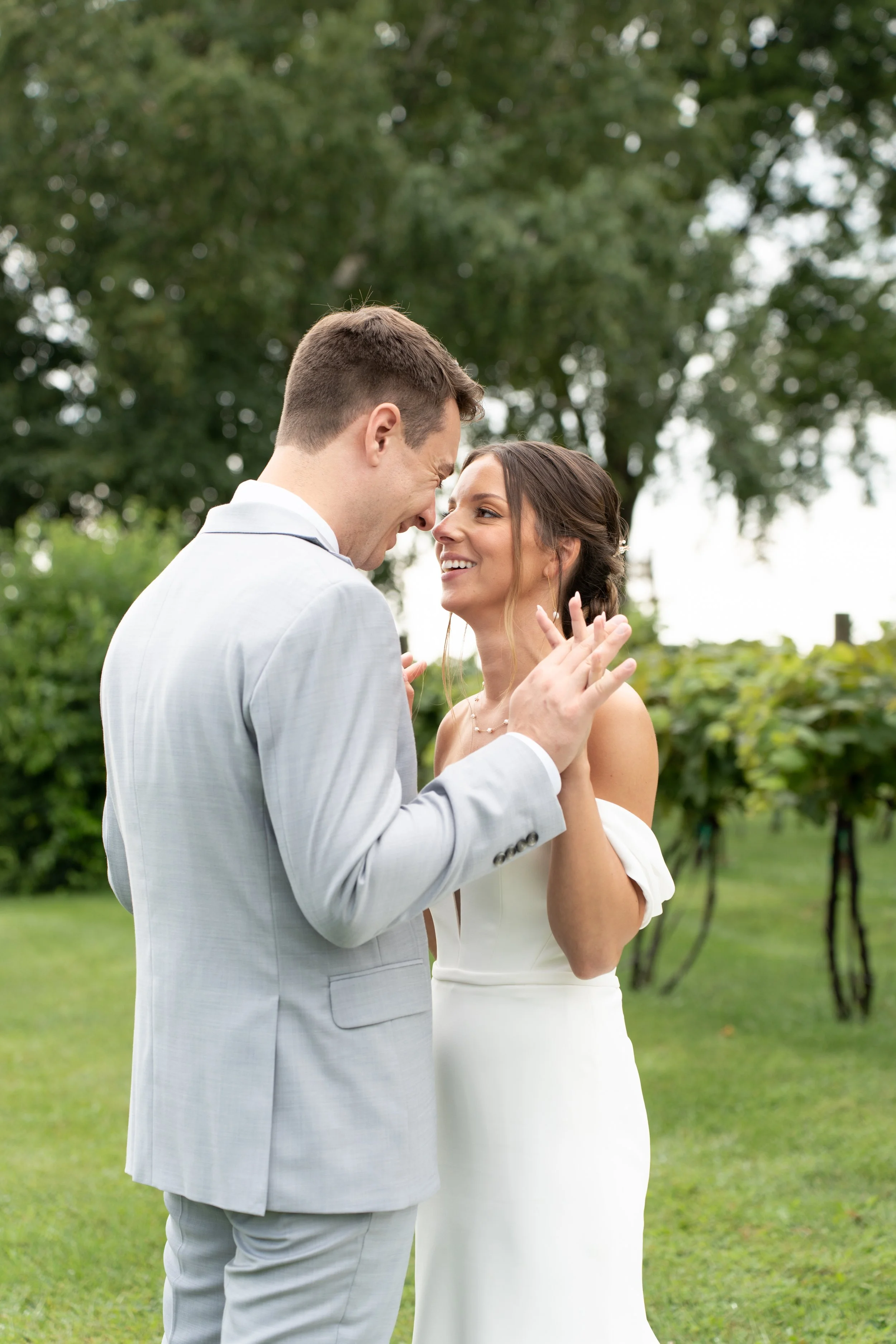 A couple in wedding attire sharing a joyful moment outdoors on a grassy lawn with green trees in the background.
