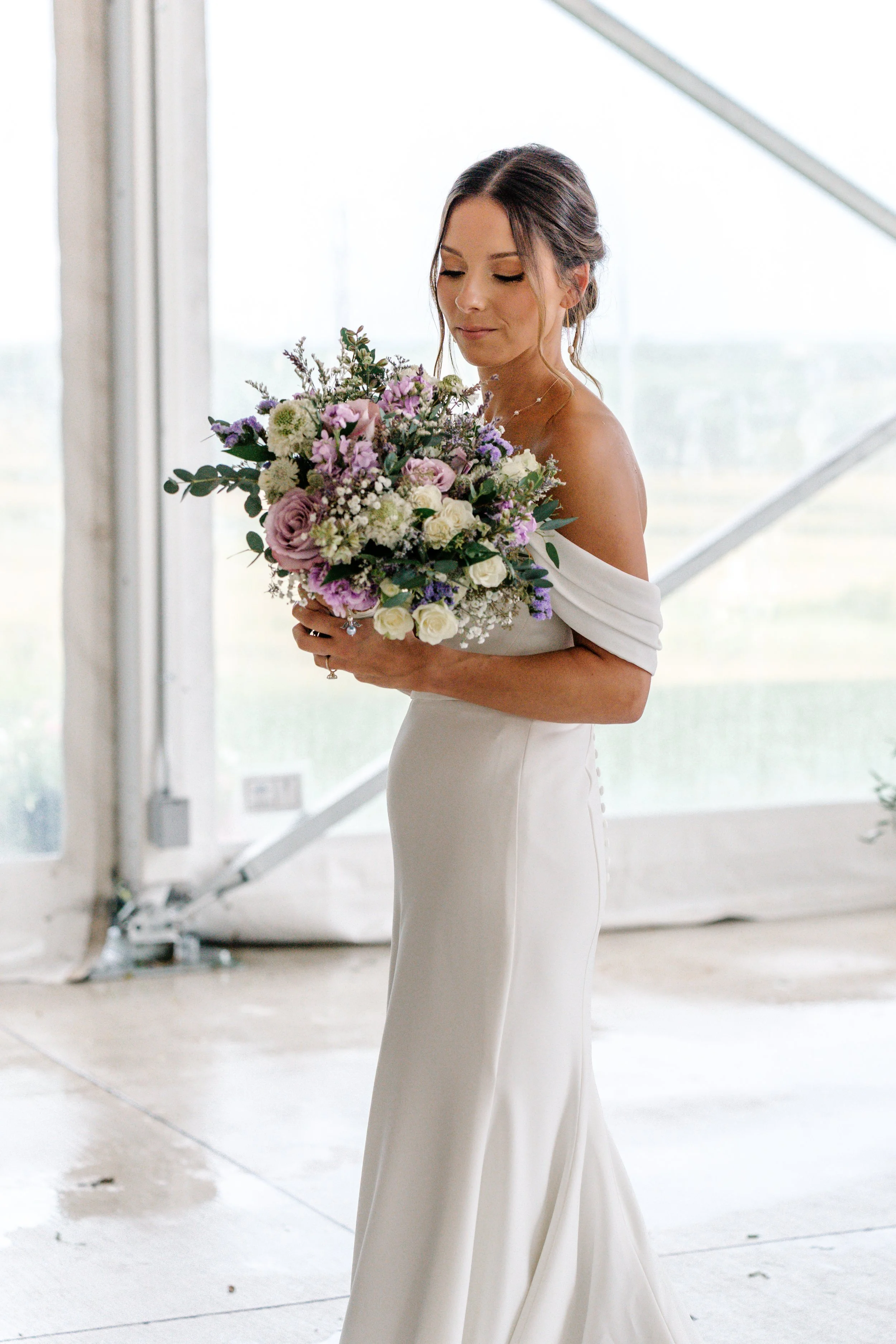 A bride in a white off-shoulder wedding dress holding a bouquet of pink, purple, and white flowers, standing indoors near large windows.
