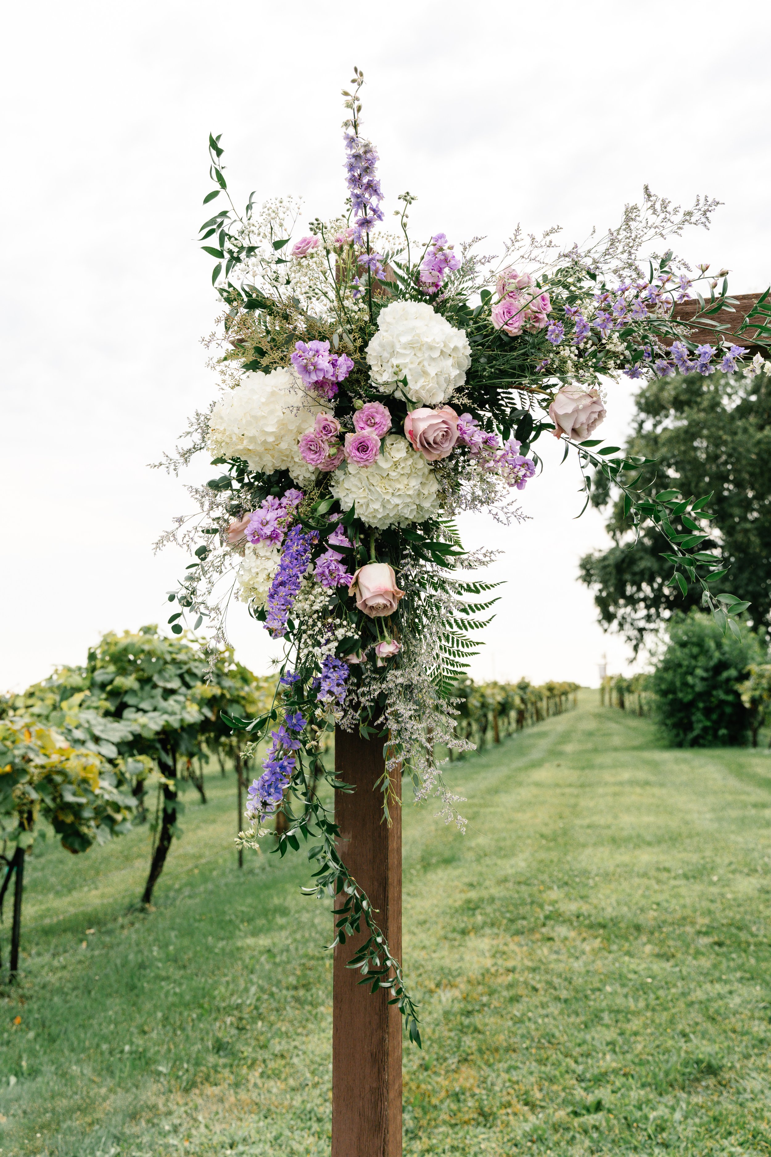 A floral arrangement with white and purple flowers on a wooden stand outdoors, with a vineyard in the background.