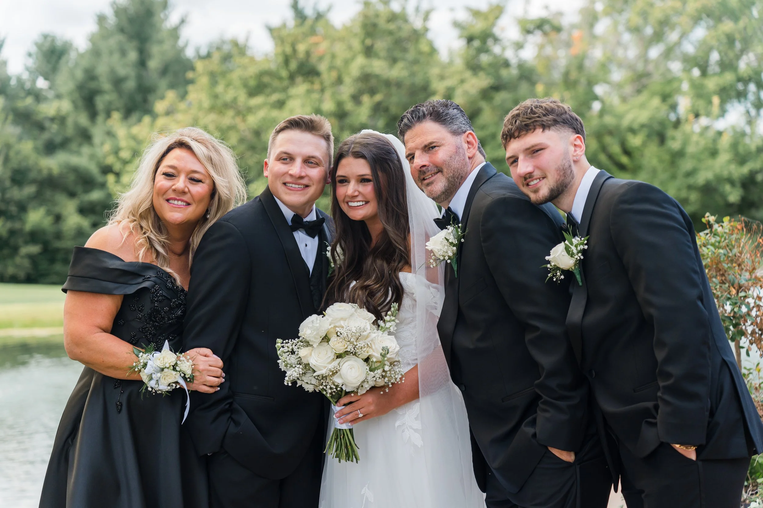 Group of five people at a wedding outdoors, with a pond and trees in the background. A bride holds a bouquet of white roses, flanked by two women and two men dressed in tuxedos. The women wear black dresses with corsages, and the men have boutonnière