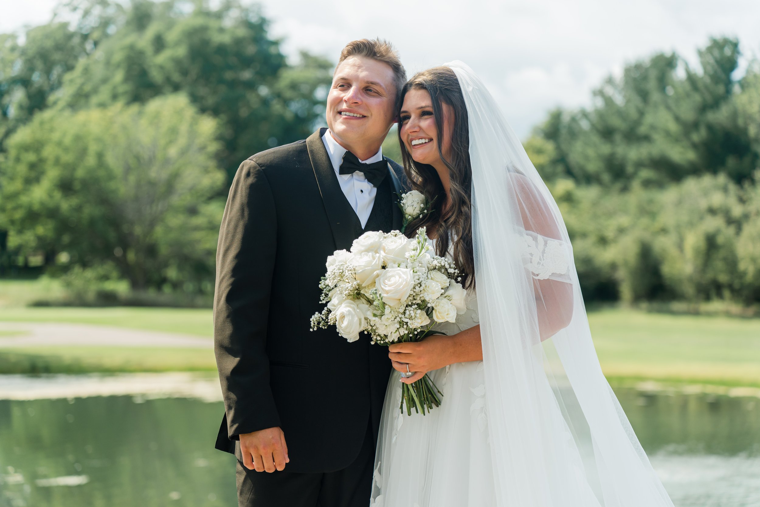 A bride and groom dressed in wedding attire standing outdoors, smiling, with greenery and a pond in the background.