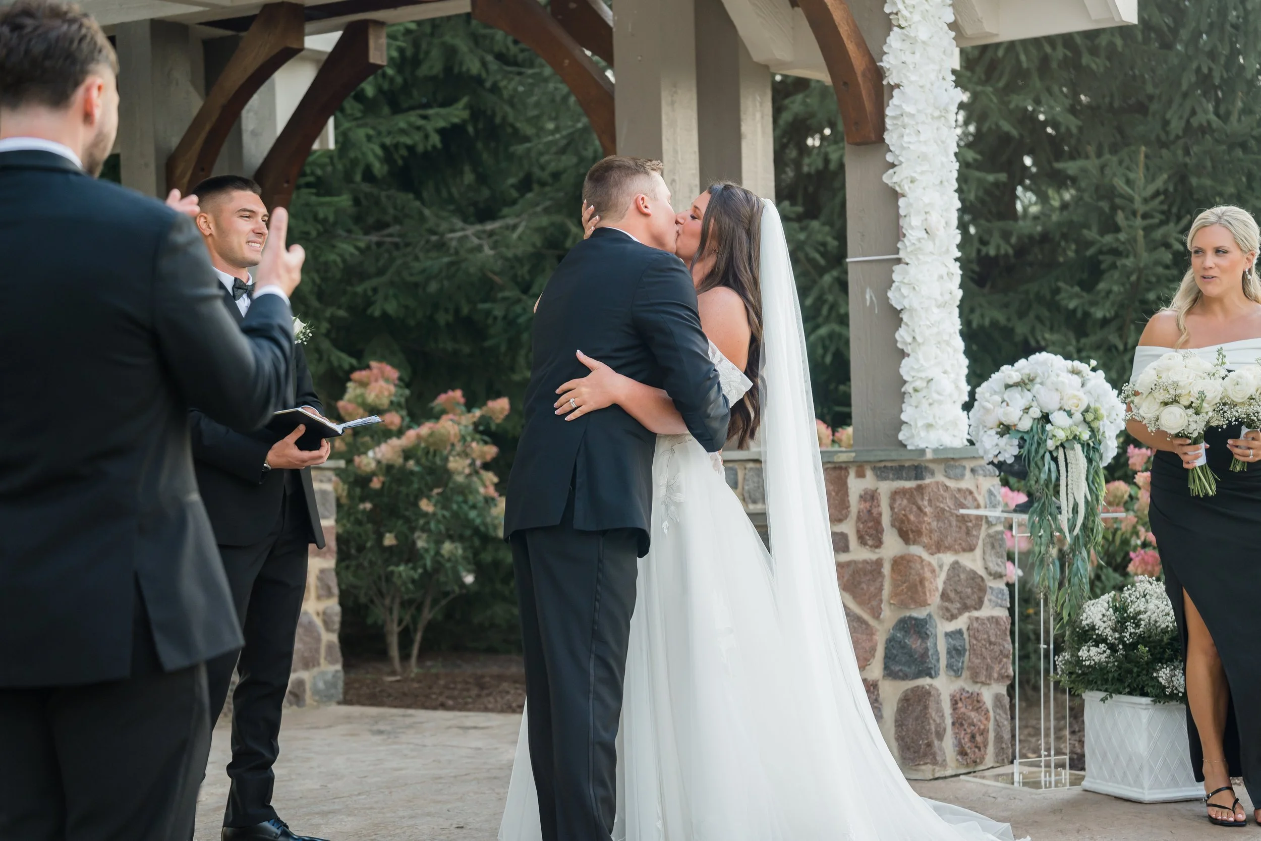 Bride and groom kissing during their wedding ceremony outdoors under a wooden structure decorated with white flowers, with bridesmaids and groomsmen standing nearby, in a garden setting.