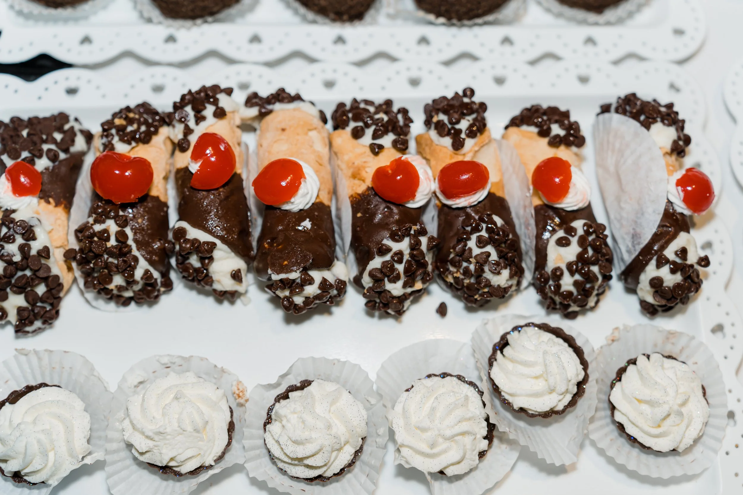 Assorted cannoli pastries with chocolate chips, whipped cream, and cherries on a white tray.
