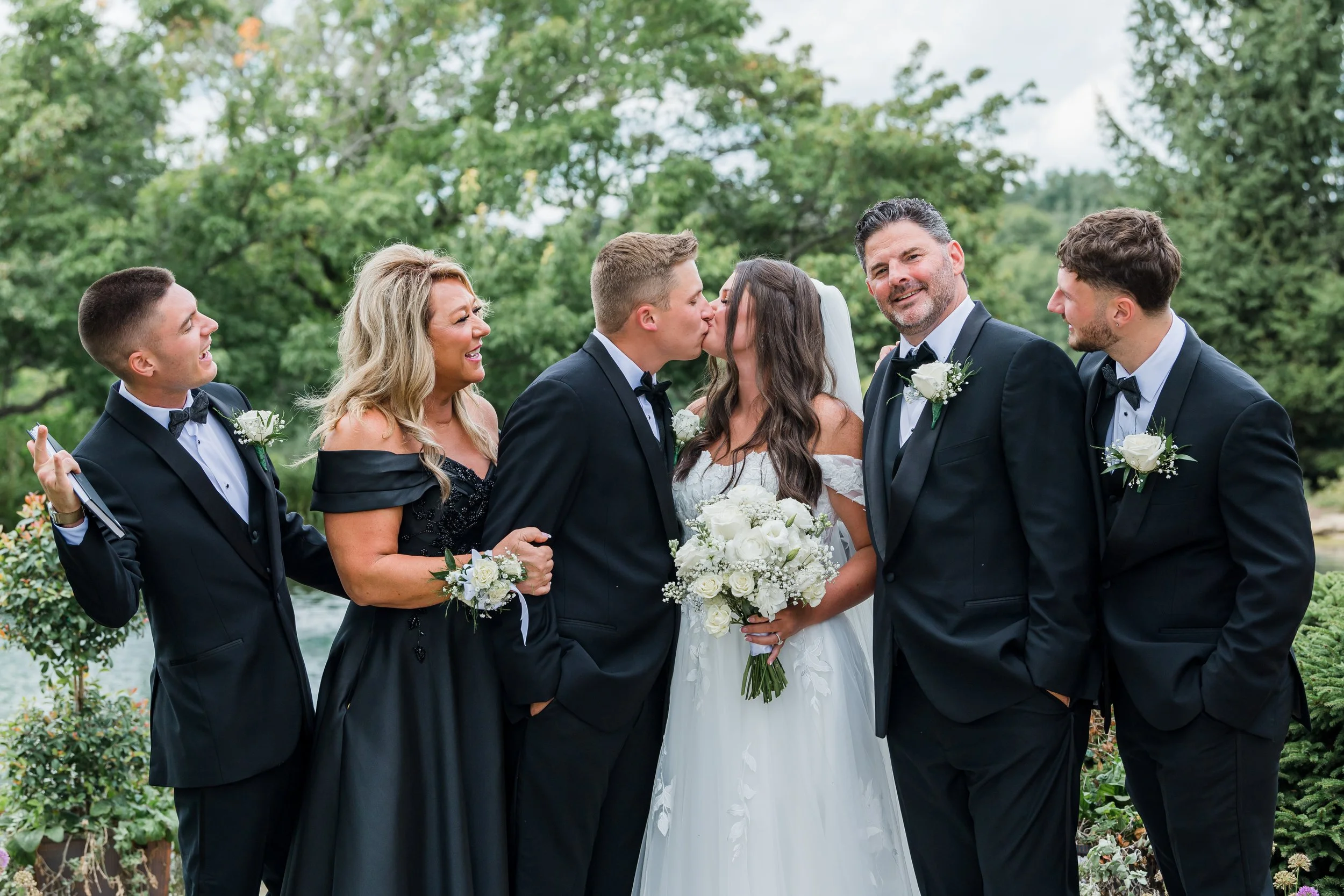 A group of six people at a wedding, with a bride and groom kissing in the center, surrounded by bridesmaids and groomsmen, outdoors with green trees in the background.