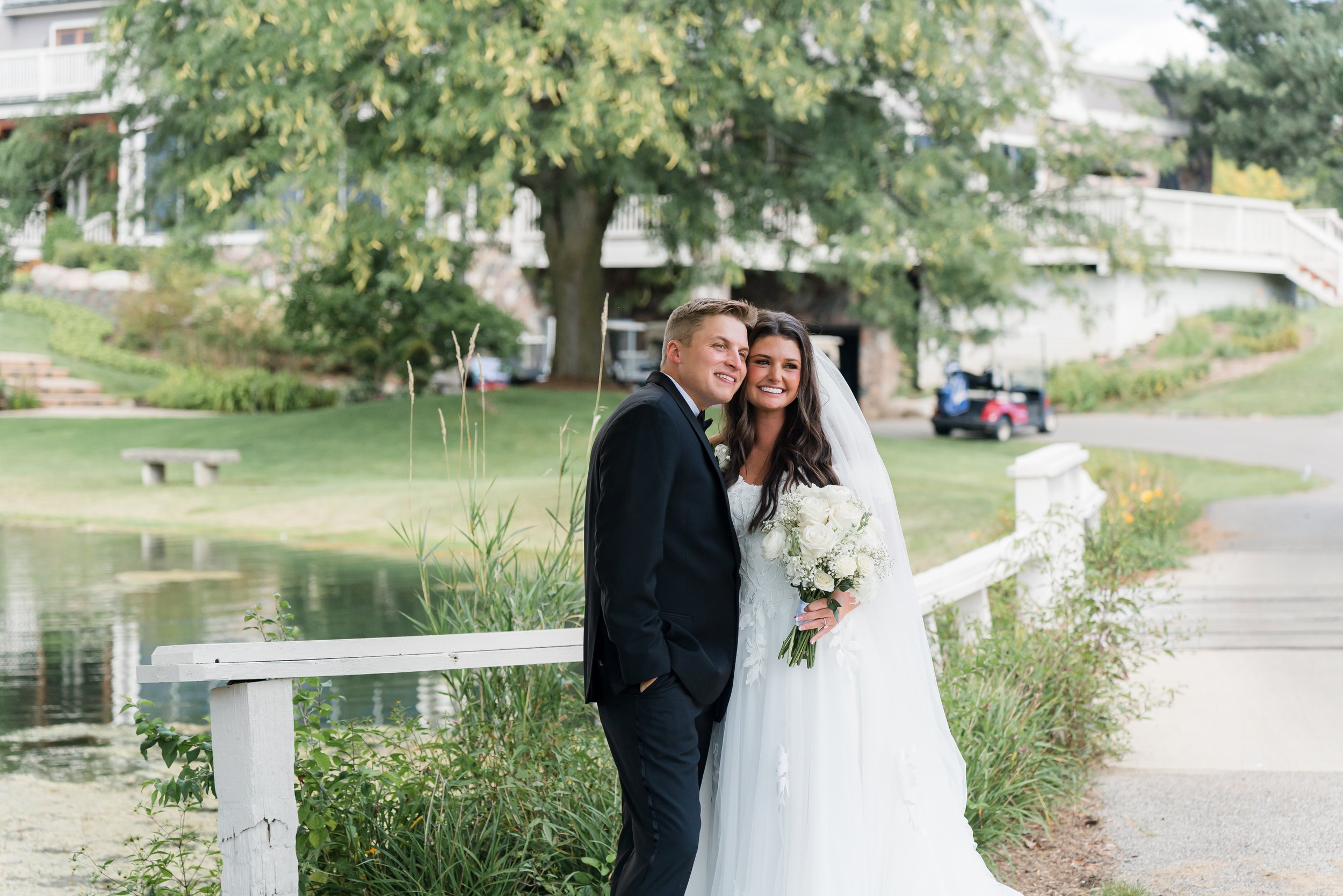 A newlywed couple smiles and poses for a photo outdoors near a pond, with greenery and a house in the background.