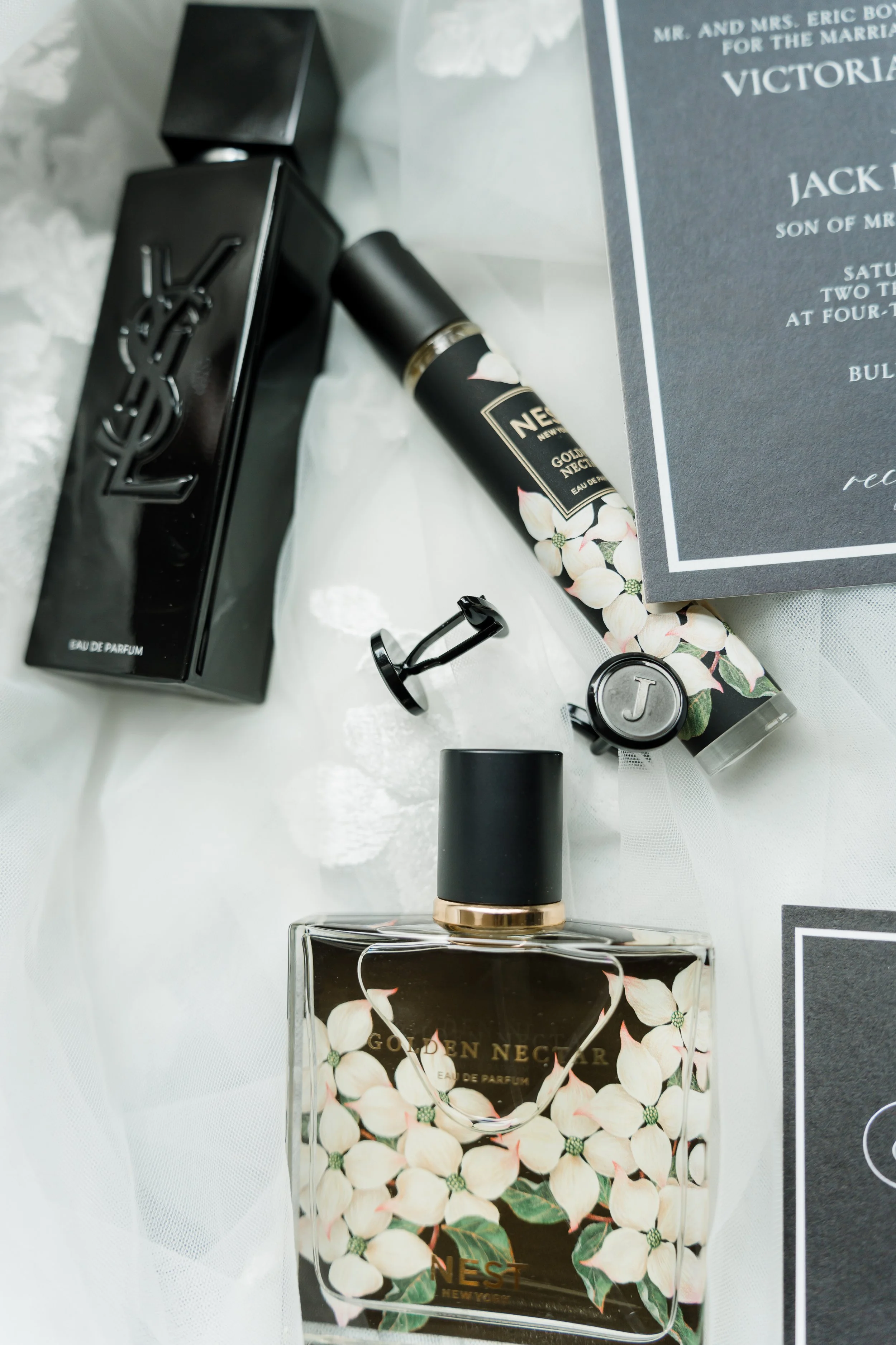 Black YSL perfume box, a bottle of Golden Nectar perfume with floral design, a roll-on perfume, a small jar with a letter J on the lid, and a black visitor badge, all arranged on white fabric and a wedding invitation.