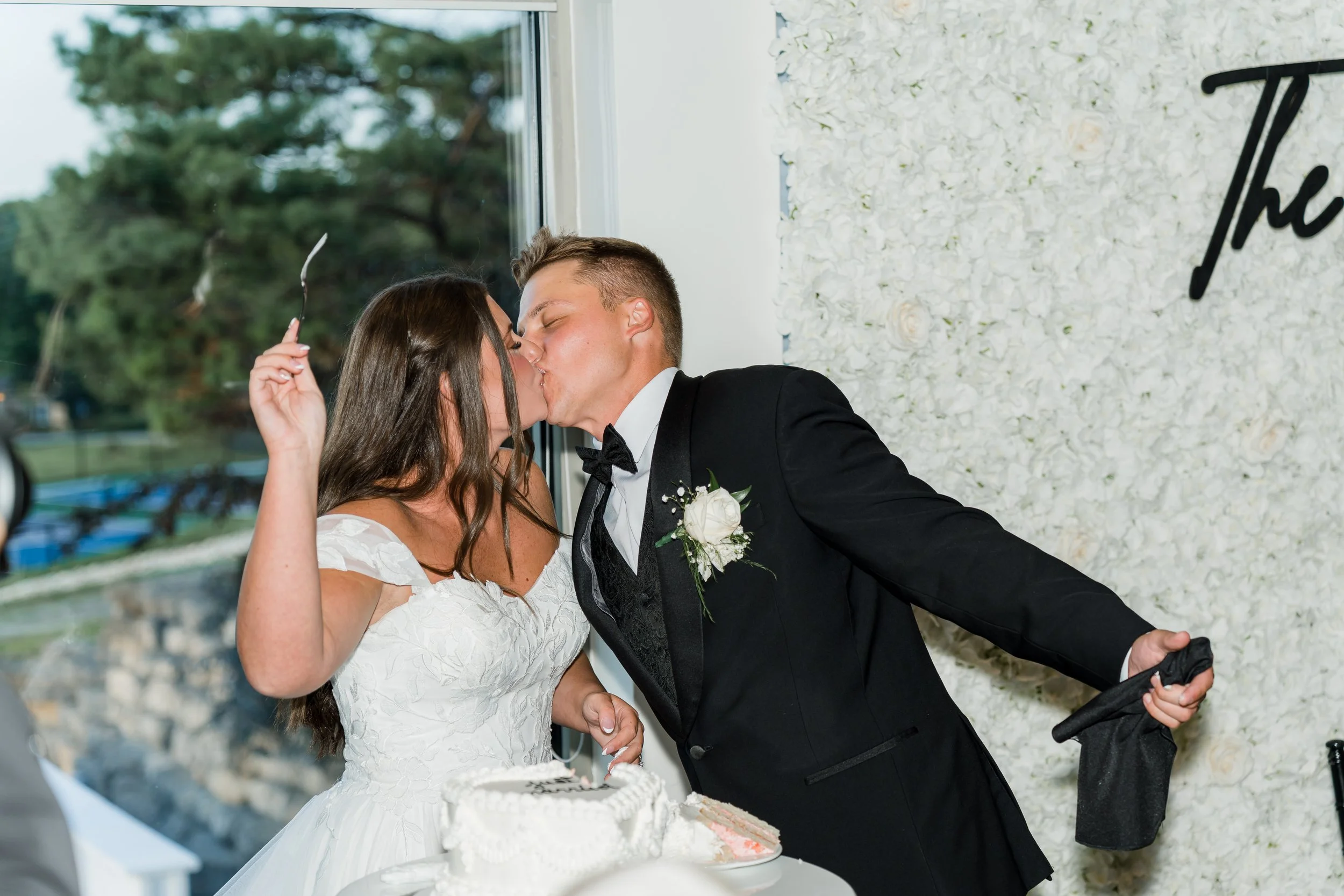 A bride and groom kissing at their wedding reception, with the bride holding a knife and the groom holding a cloth, standing in front of a white flower wall.