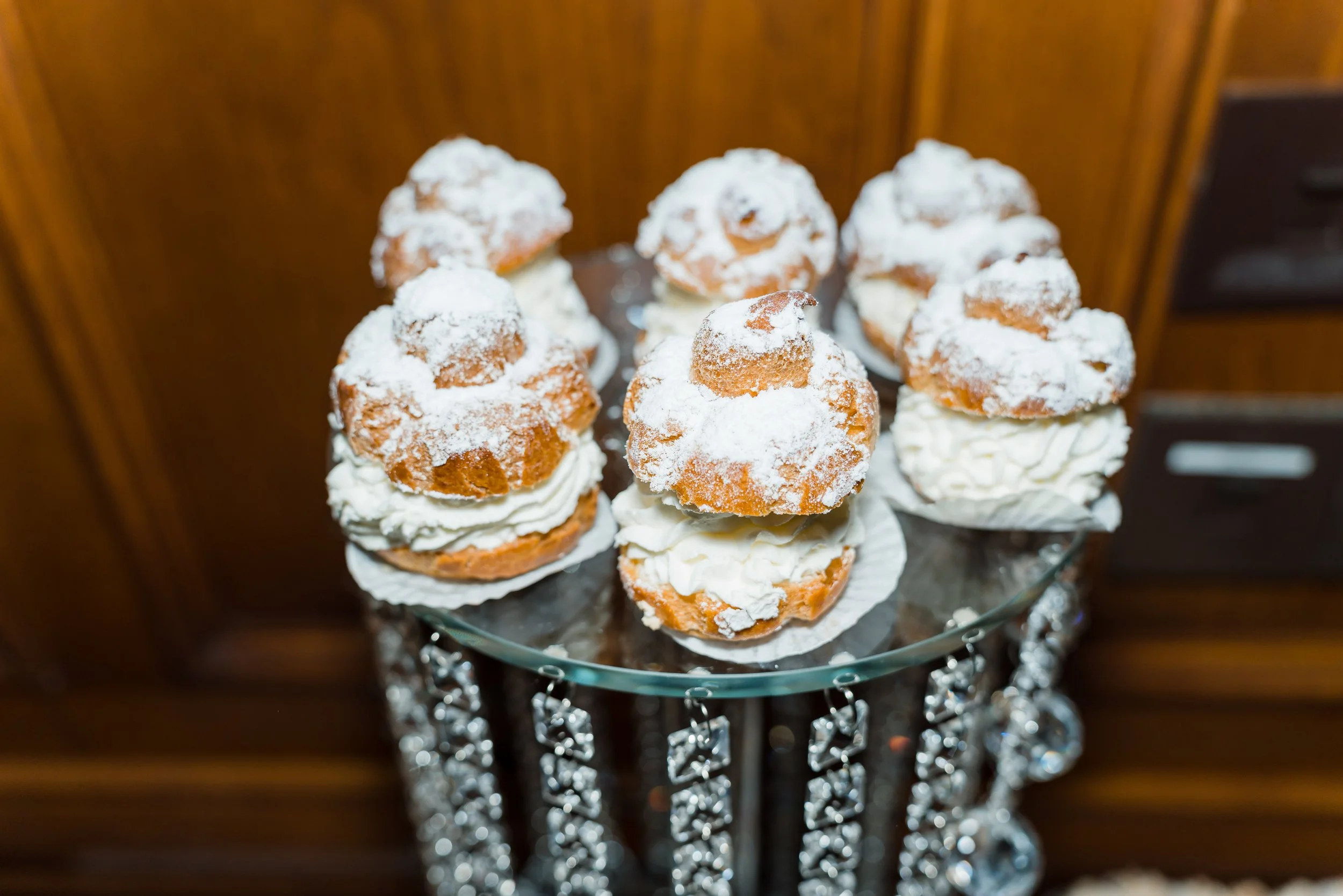 Cream puffs dusted with powdered sugar, displayed on a glass stand with decorative hanging rhinestones.