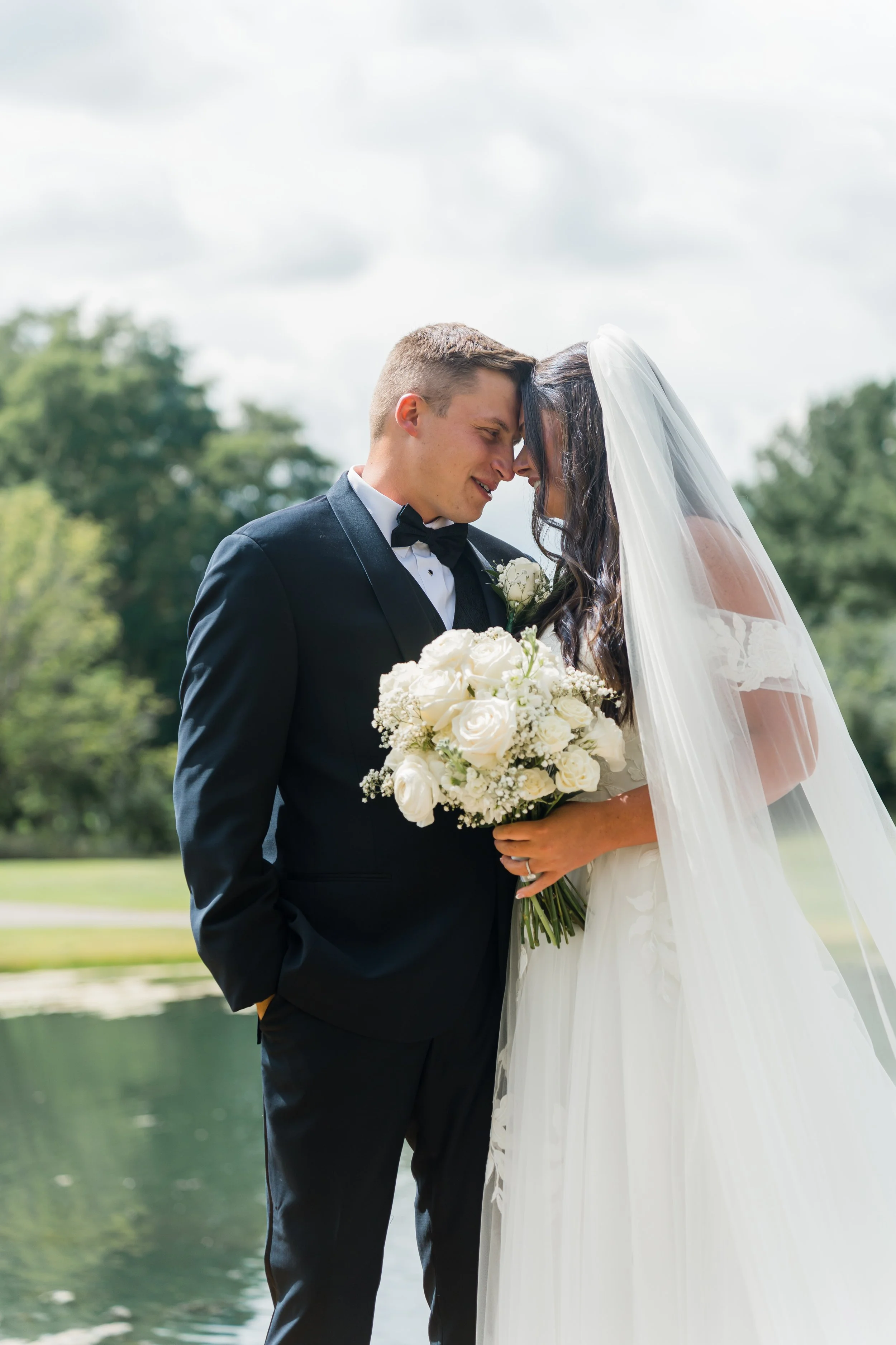 A bride and groom on their wedding day, standing close with foreheads touching, outdoors near a pond with greenery in the background. The groom is in a black tuxedo and the bride in a white wedding dress with veil, holding a bouquet of white roses.