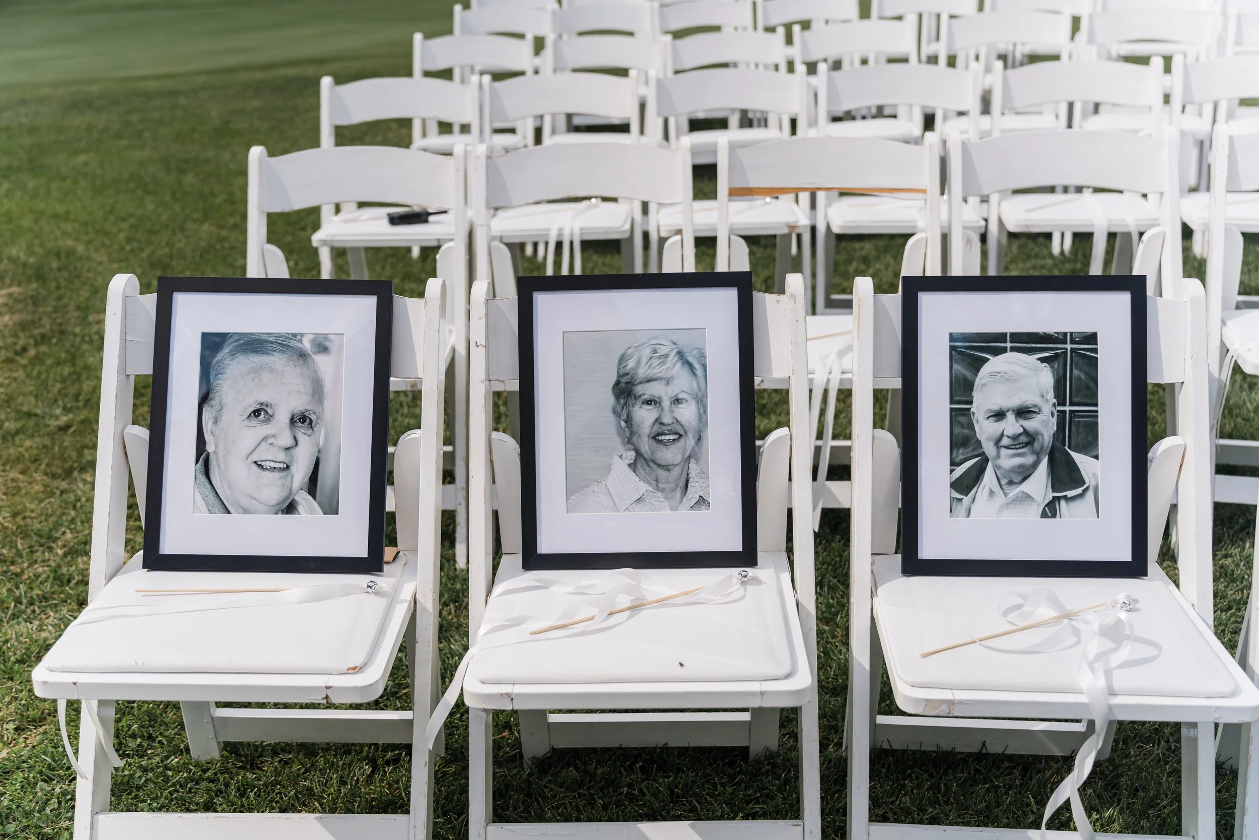 Three framed photographs of elderly individuals placed on white chairs at an outdoor memorial or ceremony, arranged on a grassy area with additional empty chairs in the background.