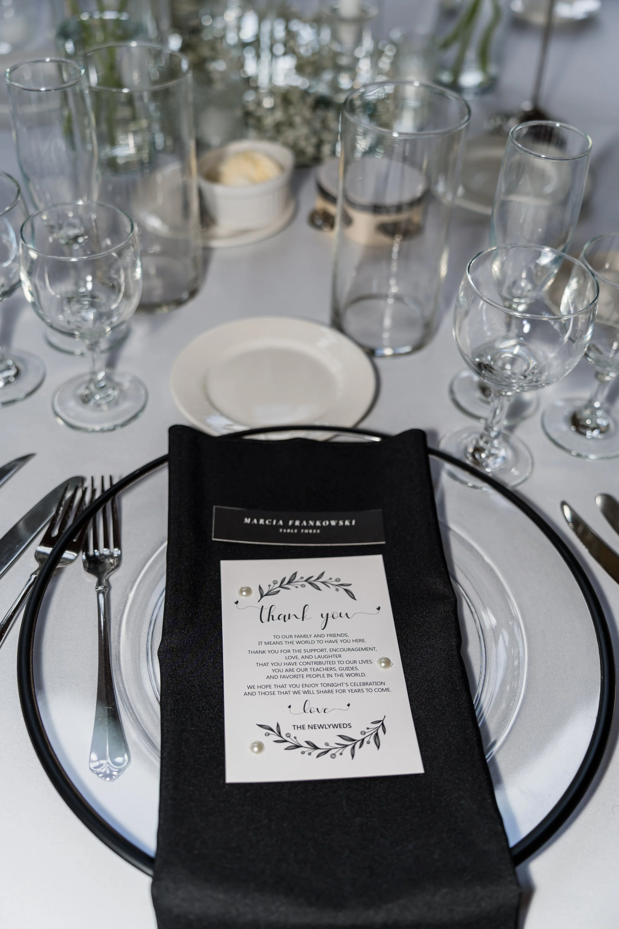 Elegant wedding reception table setting with a black napkin, a thank you card, and various glassware and silverware.