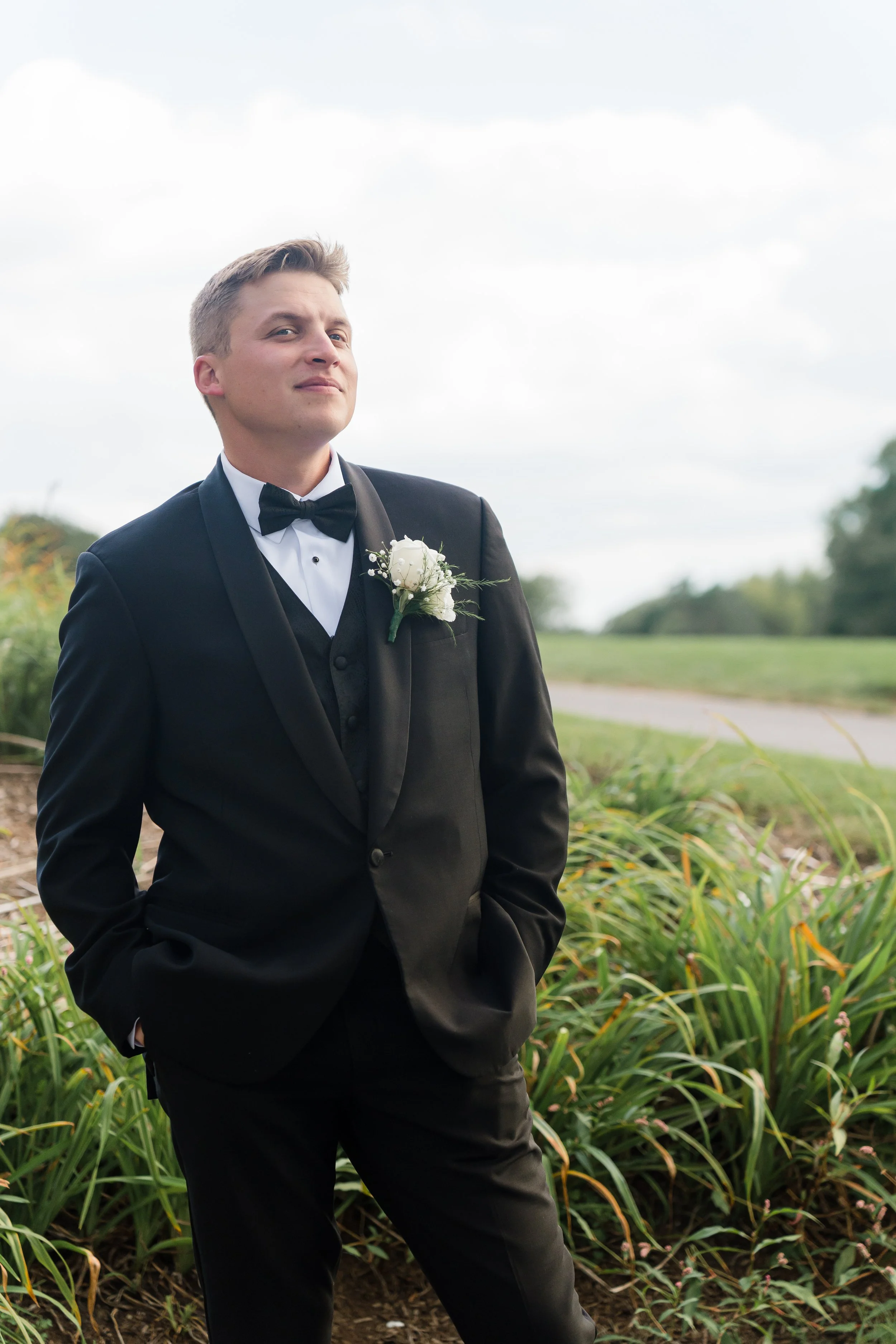 Young man in a black tuxedo with a white shirt, black bow tie, and boutonnière, standing outdoors with greenery and a cloudy sky in the background.