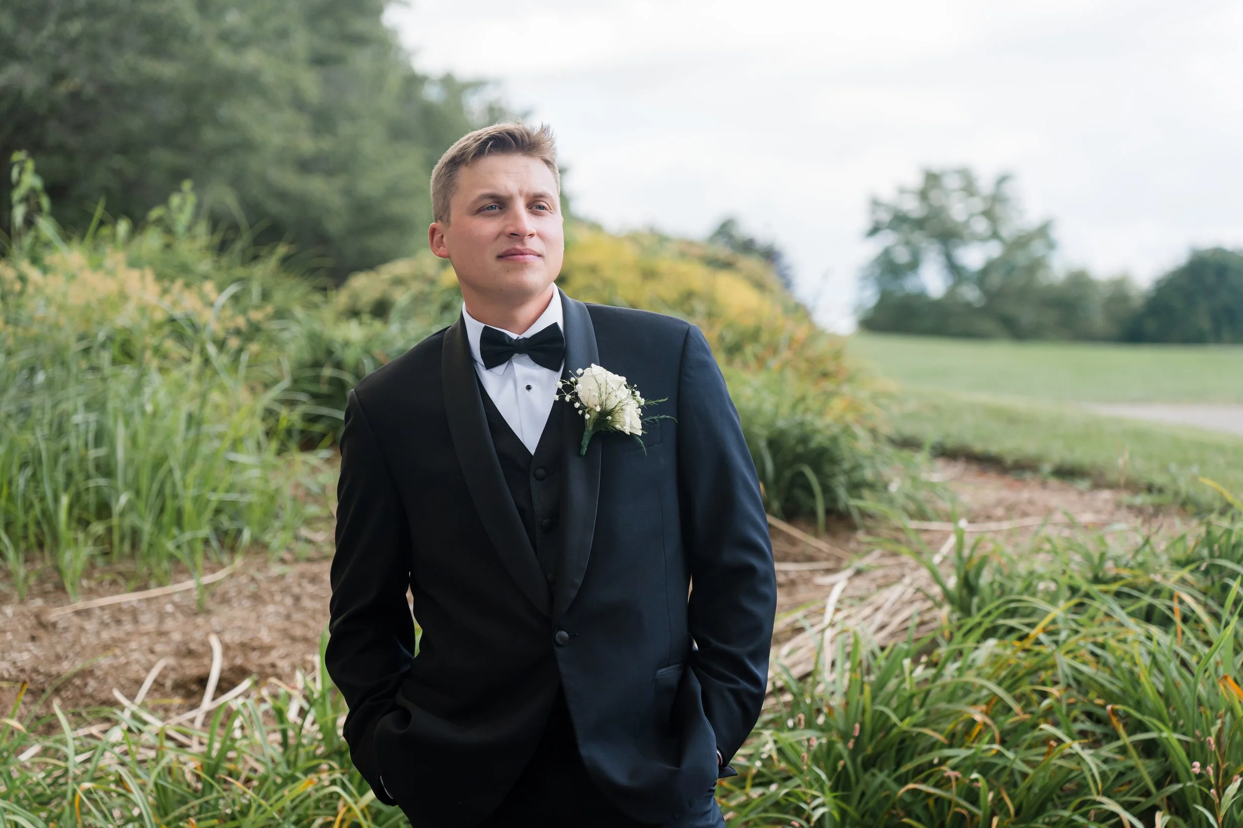 A young man in a tuxedo with a white boutonniere walks outdoors in a garden or park, with grass, bushes, and trees in the background.
