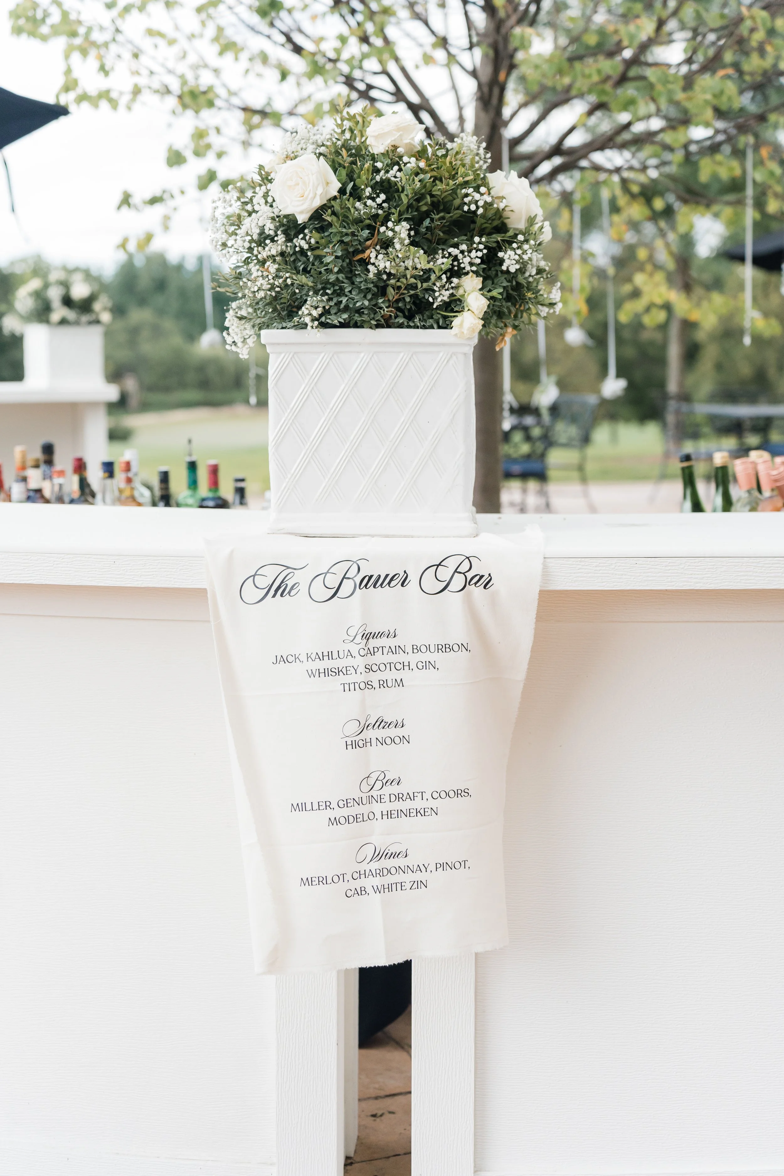 A white floral arrangement in a white basket on a bar with a sign displaying drink options for a wedding or event.