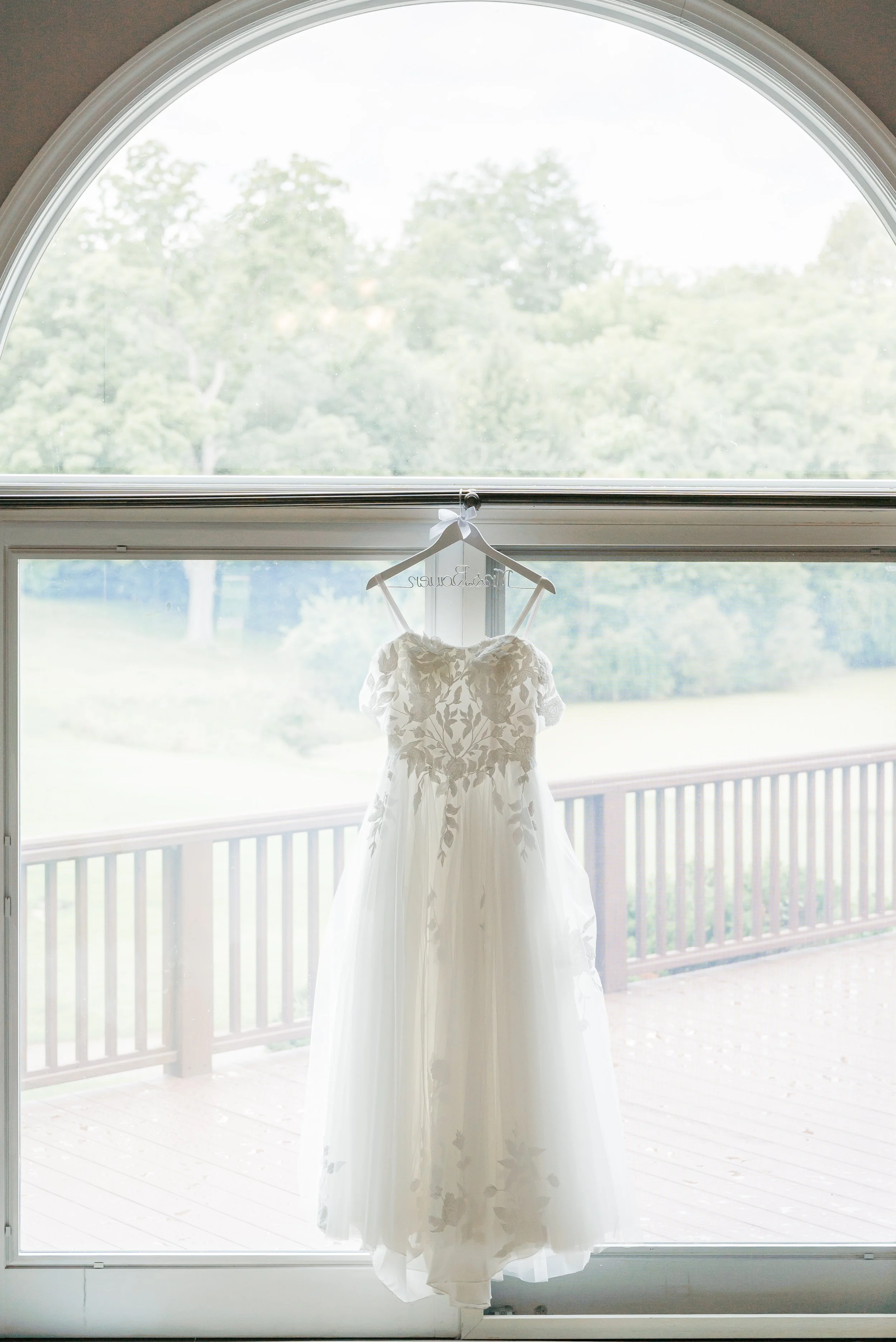 Wedding dress hanging in front of a large window with a view of trees and a deck outside.