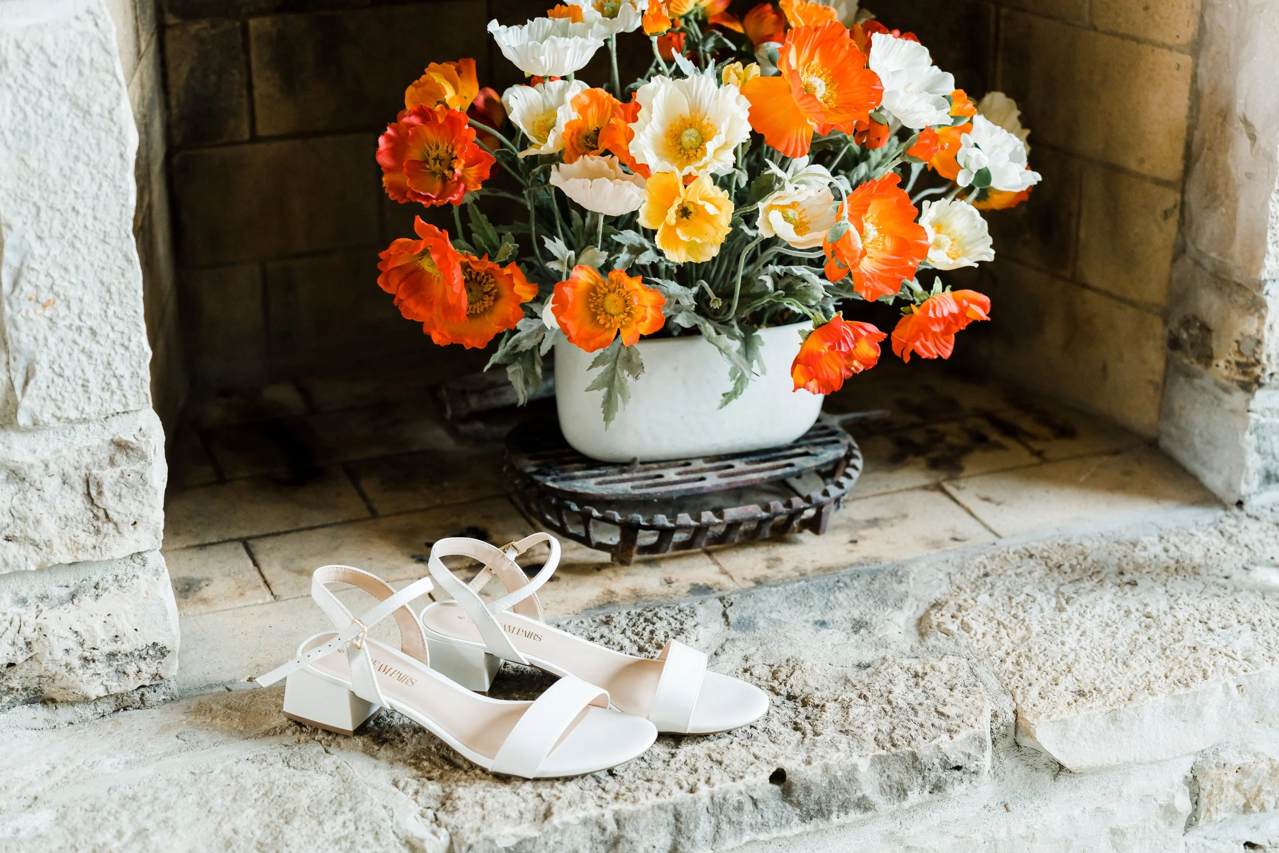 A pair of white heeled sandals placed on a stone hearth in front of a fireplace, with a white vase of orange and white poppy flowers on a black metal stand inside the fireplace.