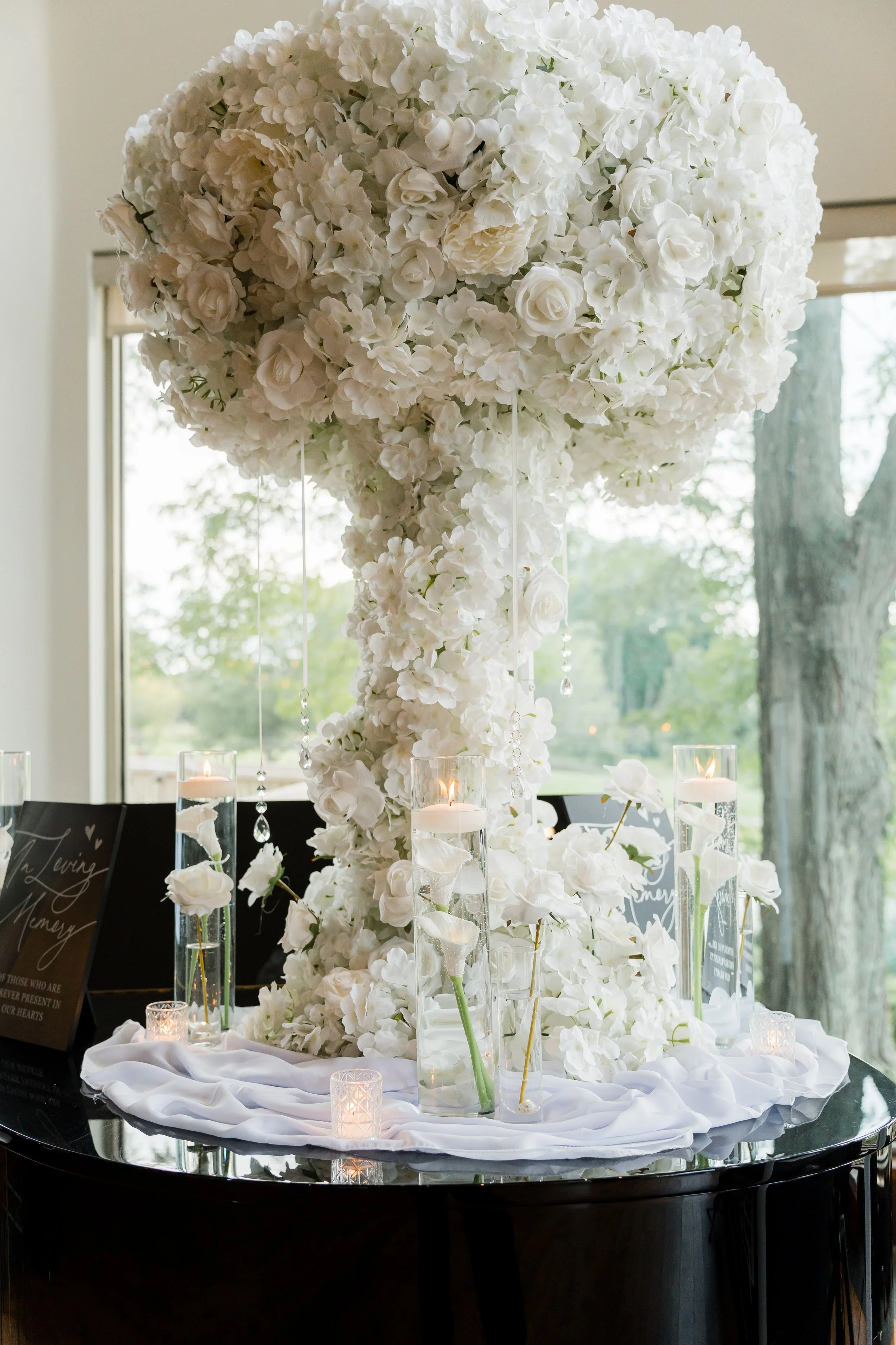 A large floral arrangement of white flowers on a piano, decorated with candles and small signs, likely for a wedding or memorial.