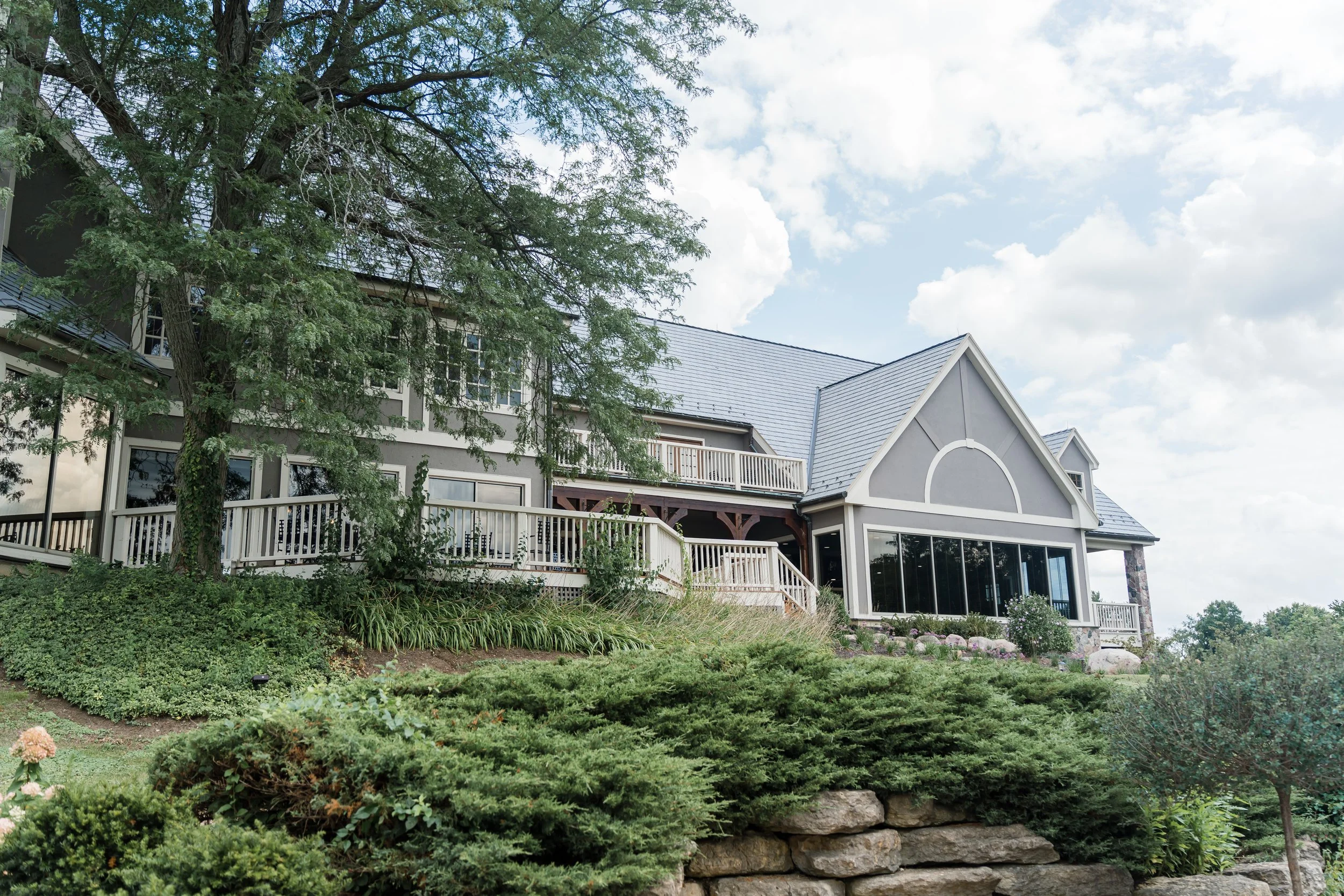 A large house on a hillside with multiple balconies, large windows, and a gray roof, surrounded by green trees and bushes under a partly cloudy sky.