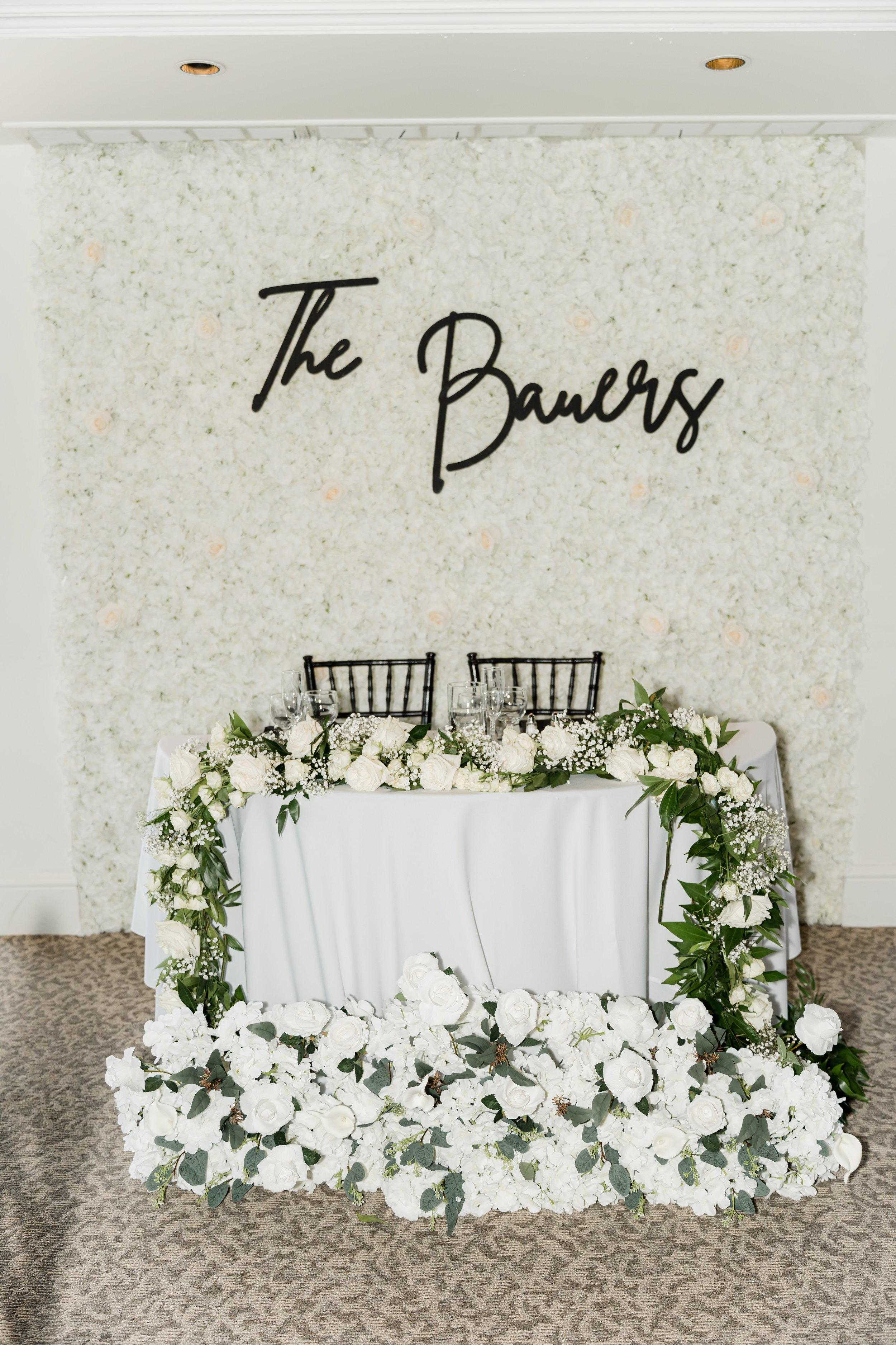 Elegant wedding or event setup with a floral backdrop, a table decorated with white flowers and greenery, two black chairs, and a large sign reading 'The Bowers' on the floral wall.