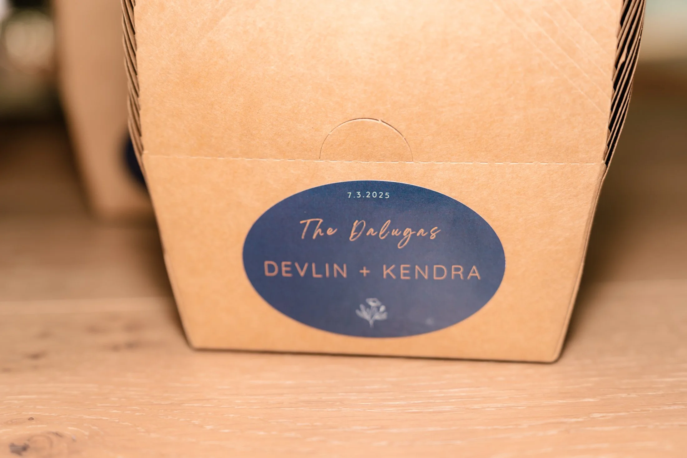 A brown cardboard box with a dark blue oval label that says "The Dalugas DEVLIN + KENDRA" and the date "7.3.2025" on top, placed on a wooden surface.