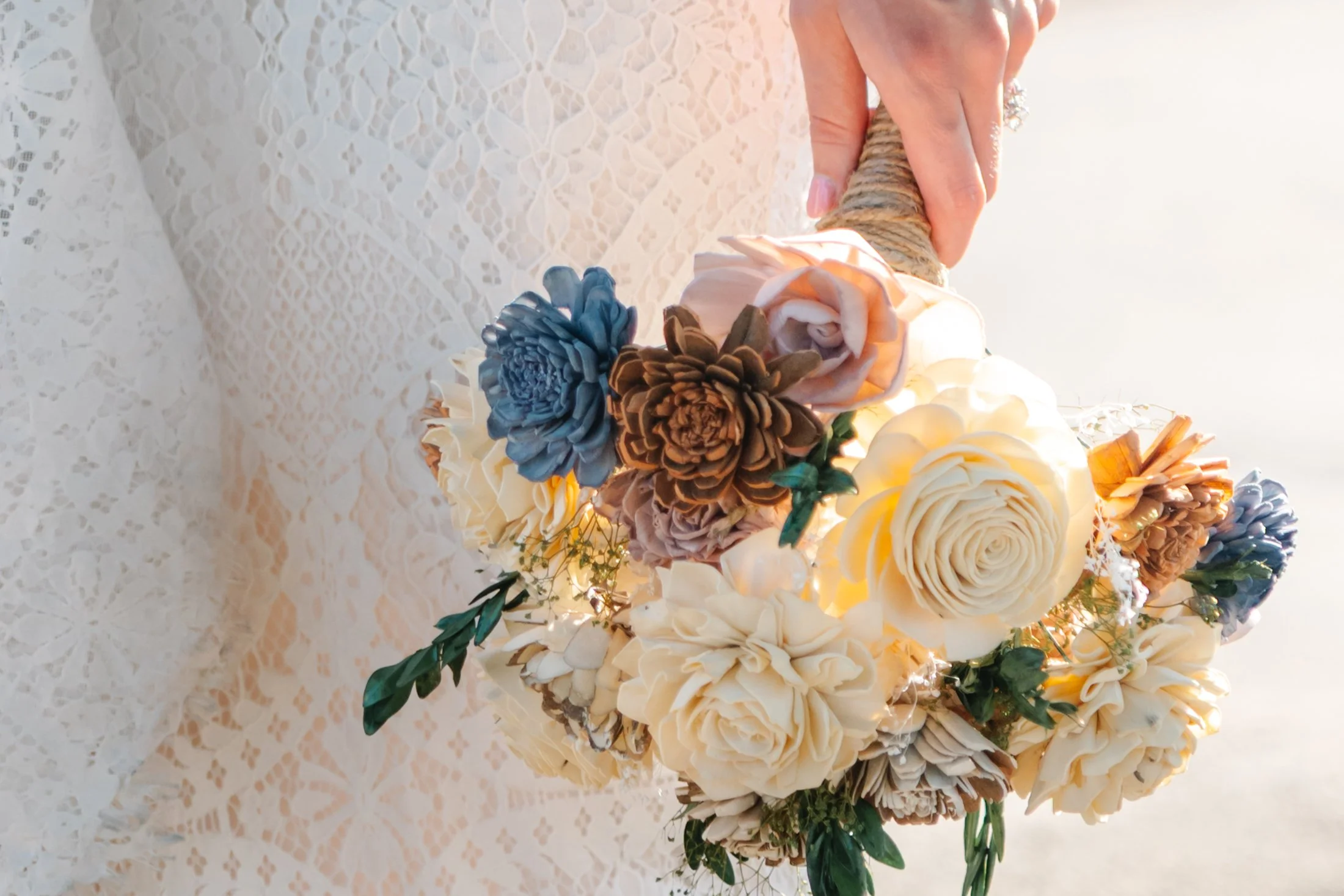 A person holding a bouquet of artificial flowers, including cream, brown, blue, and pink roses, against a lace-covered background.
