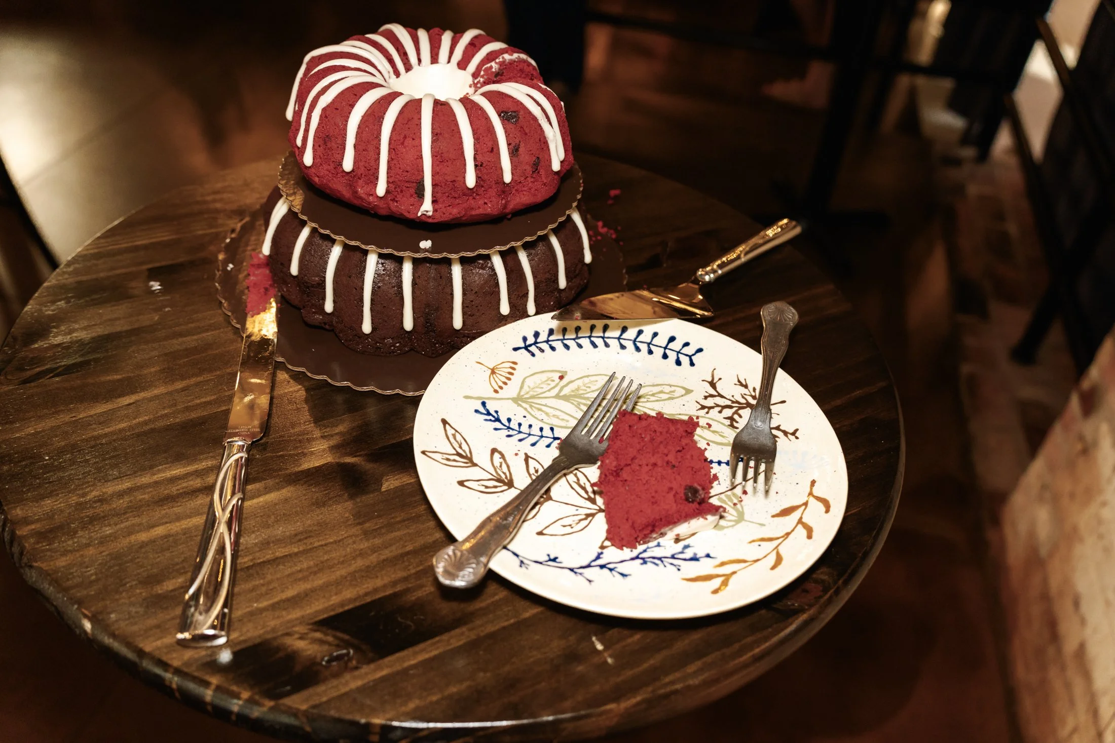 A red velvet cake with white icing drizzle, topped with a red Bundt cake and a chocolate disc, on a wooden table with a plate with a slice of cake, two forks, and a cake knife.