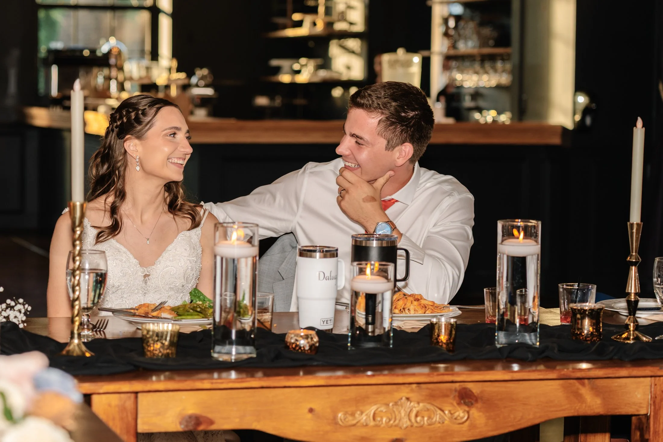 A bride and groom sitting at a wedding reception table, smiling and looking at each other, surrounded by lit candles and table decorations.