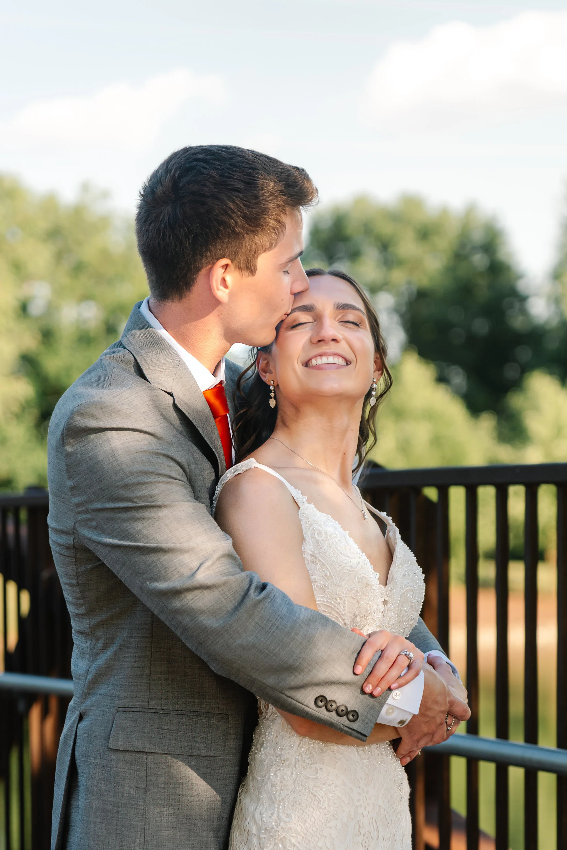 A groom kissing a smiling bride on the forehead outdoors on a sunny day.