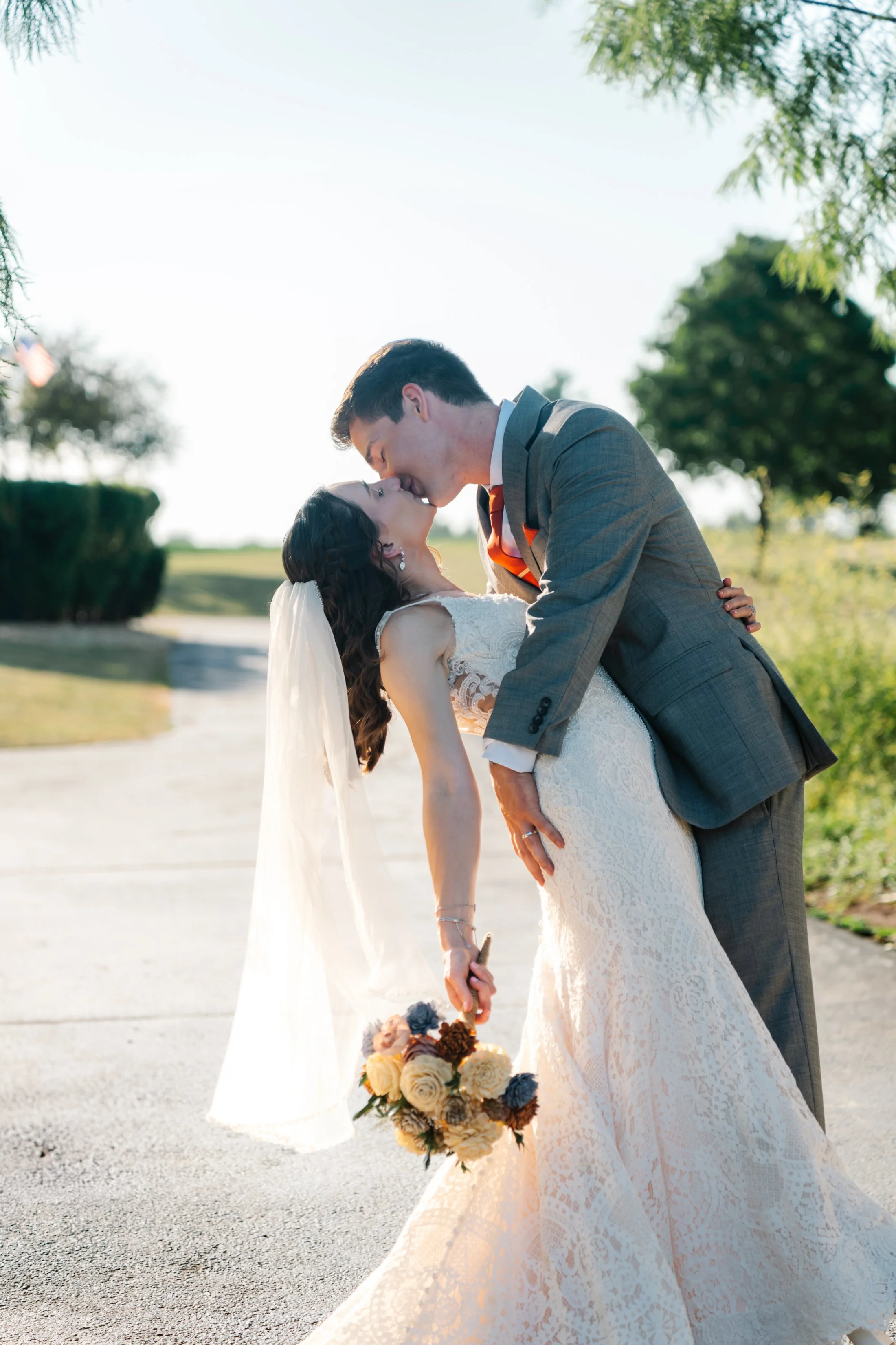 A wedding couple sharing a kiss outdoors, with the bride in a white lace gown holding a bouquet and the groom in a gray suit, sunlight illuminating the scene.