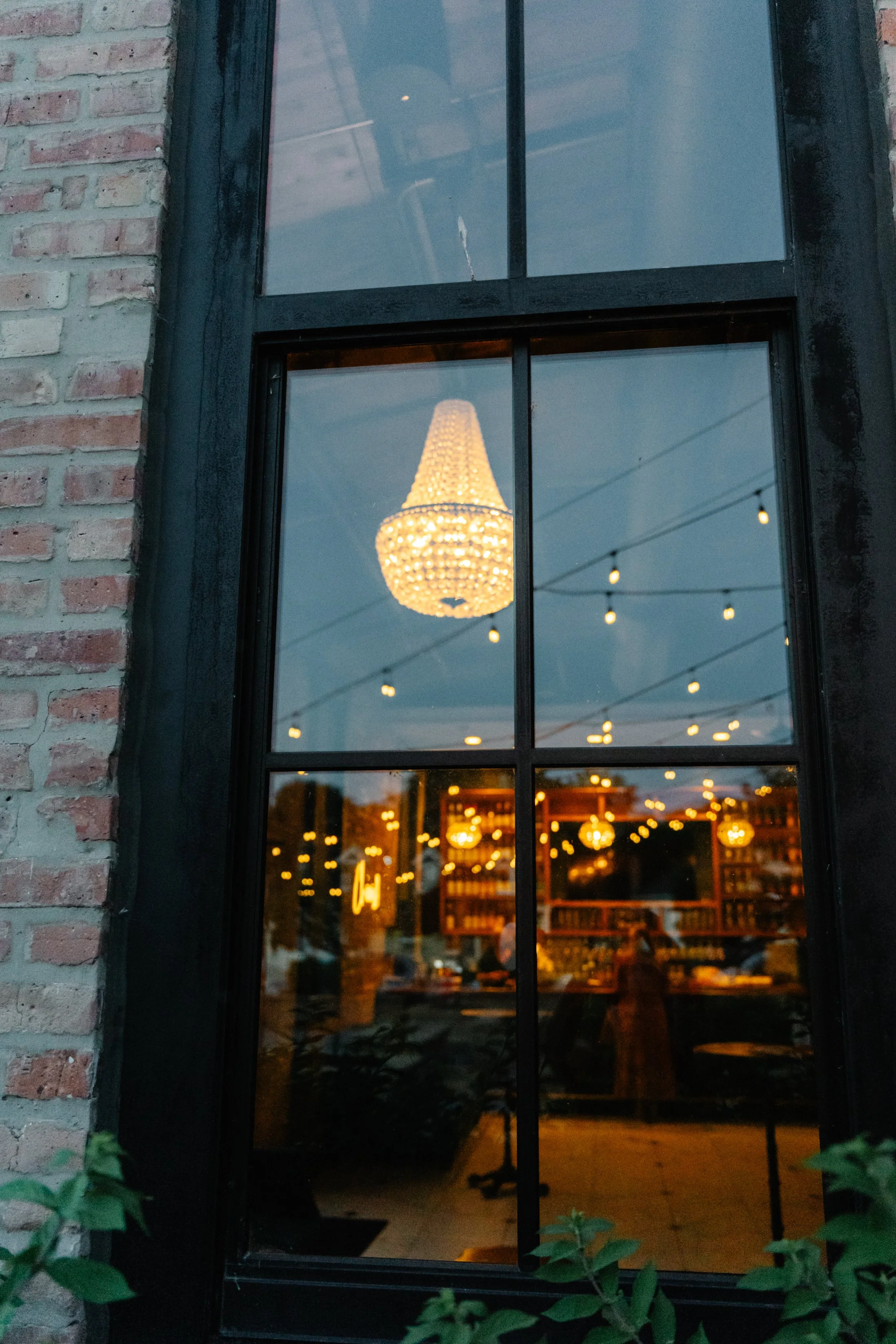 View through a large black-framed window showing a hanging chandelier inside a cozy space with string lights overhead, brick exterior wall on the side, and some greenery at the bottom.
