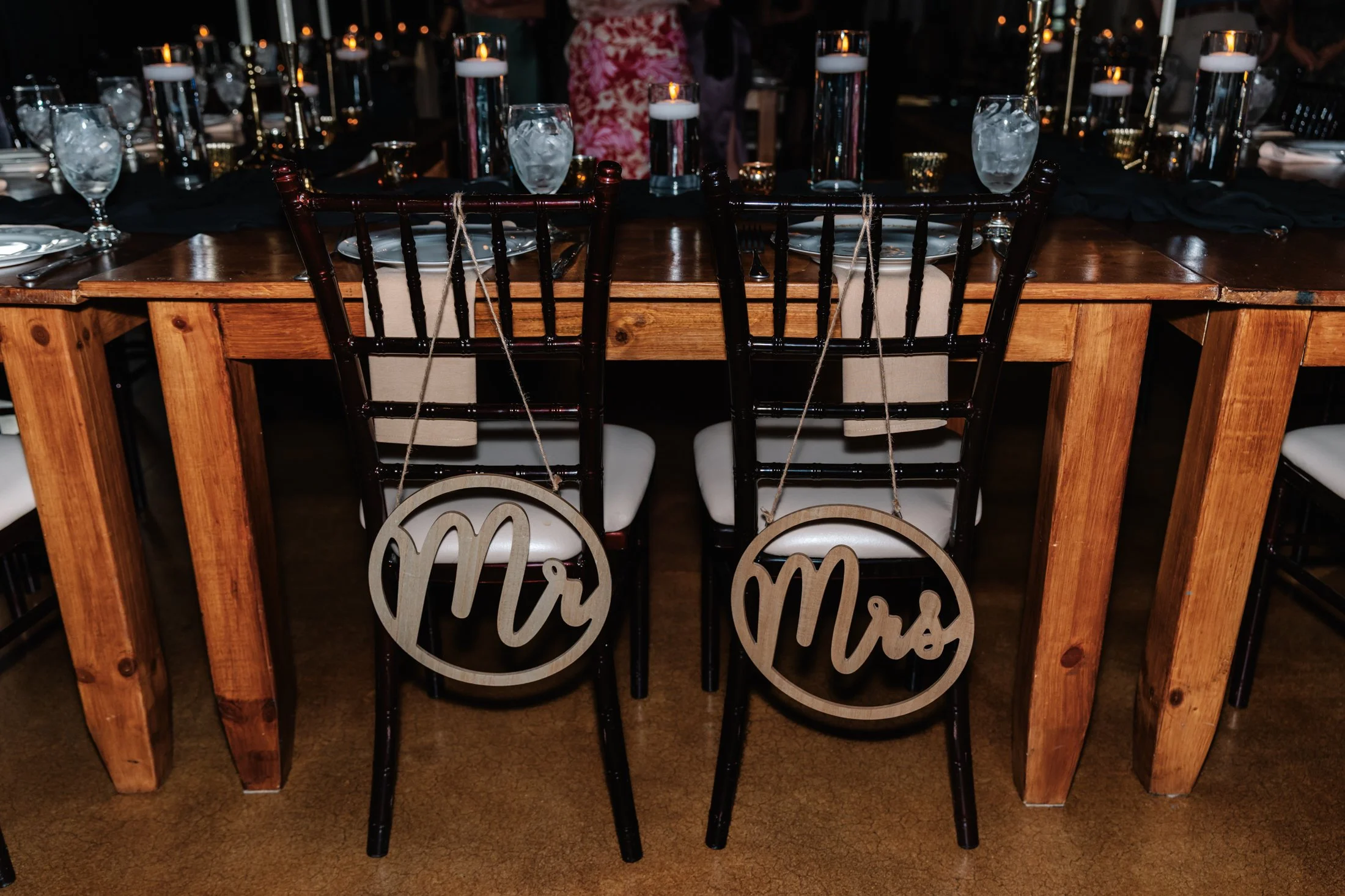 Wedding reception table with black and white decorations, glassware, and candles. Two wooden chairs with 'Mr' and 'Mrs' signs hanging on the back.