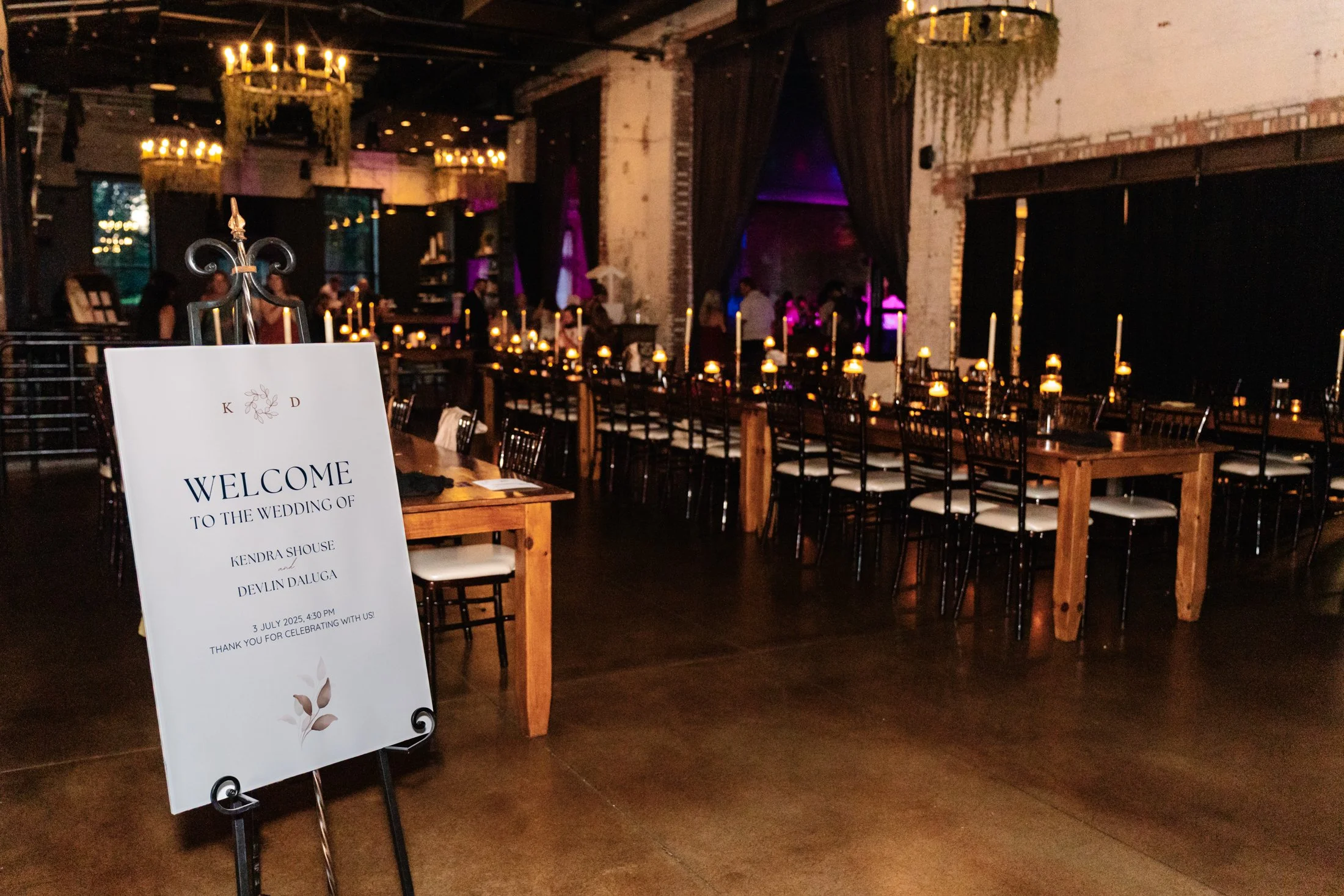 A wedding reception venue with a long rectangular wooden table surrounded by black chairs with white cushions, decorated with lit candles in glass holders. In the foreground, a welcome sign on an easel announces the wedding of Kendra Shouse and Devli