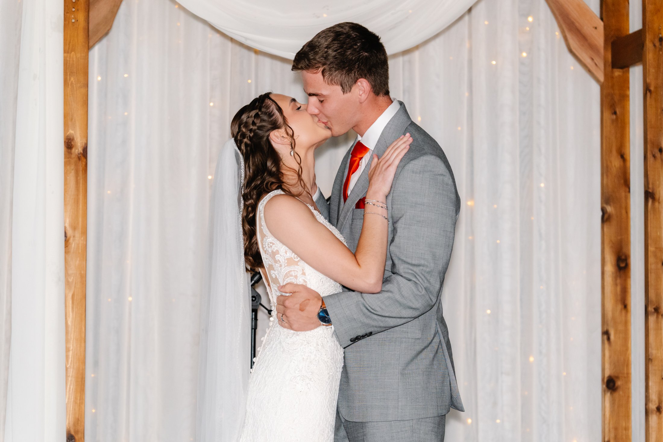 A bride and groom sharing a kiss on their wedding day, standing under a decorated arch with white drapes and string lights in the background.