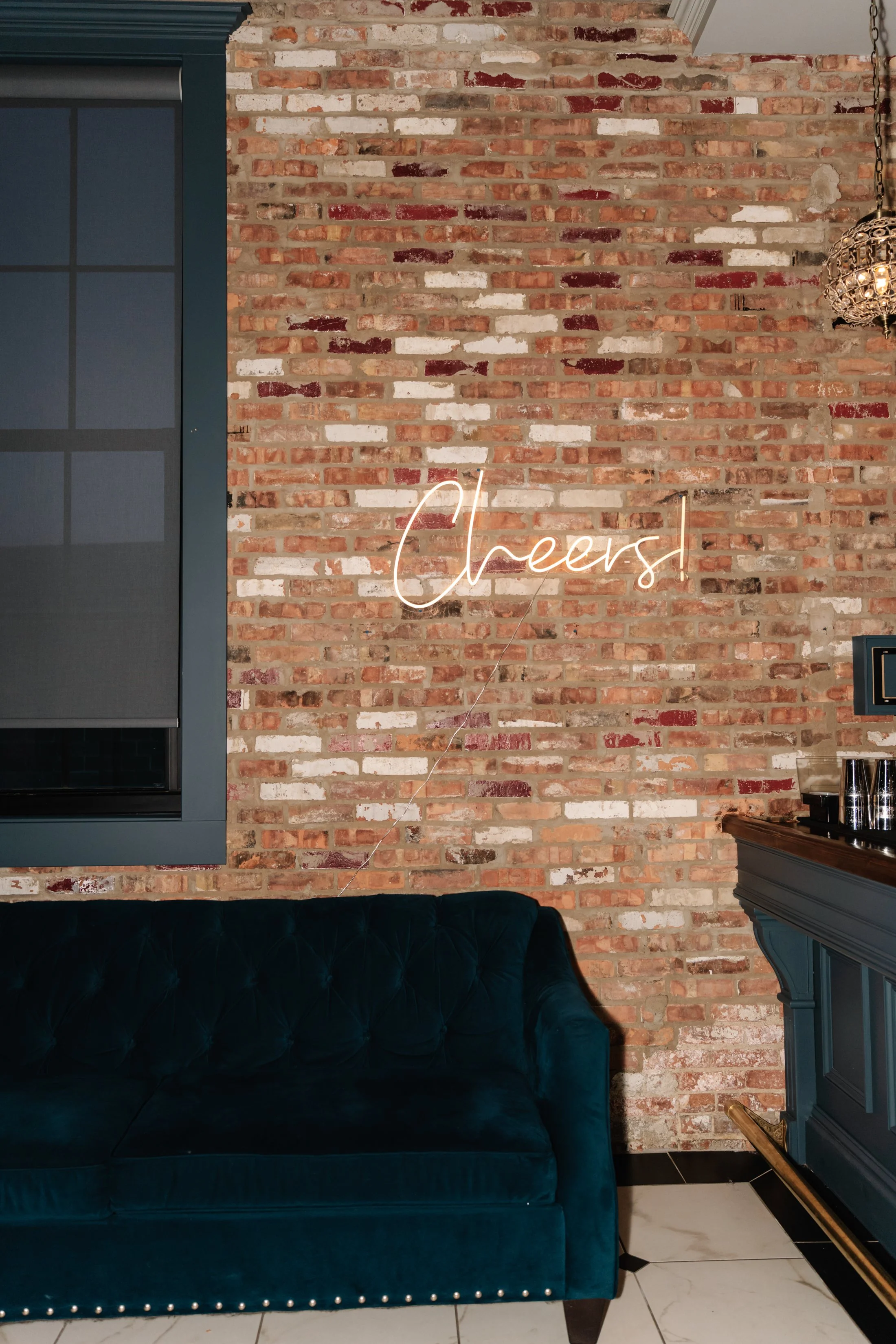 Neon sign on brick wall that reads 'Cheers!', with a dark green velvet sofa and black and white tiled floor.