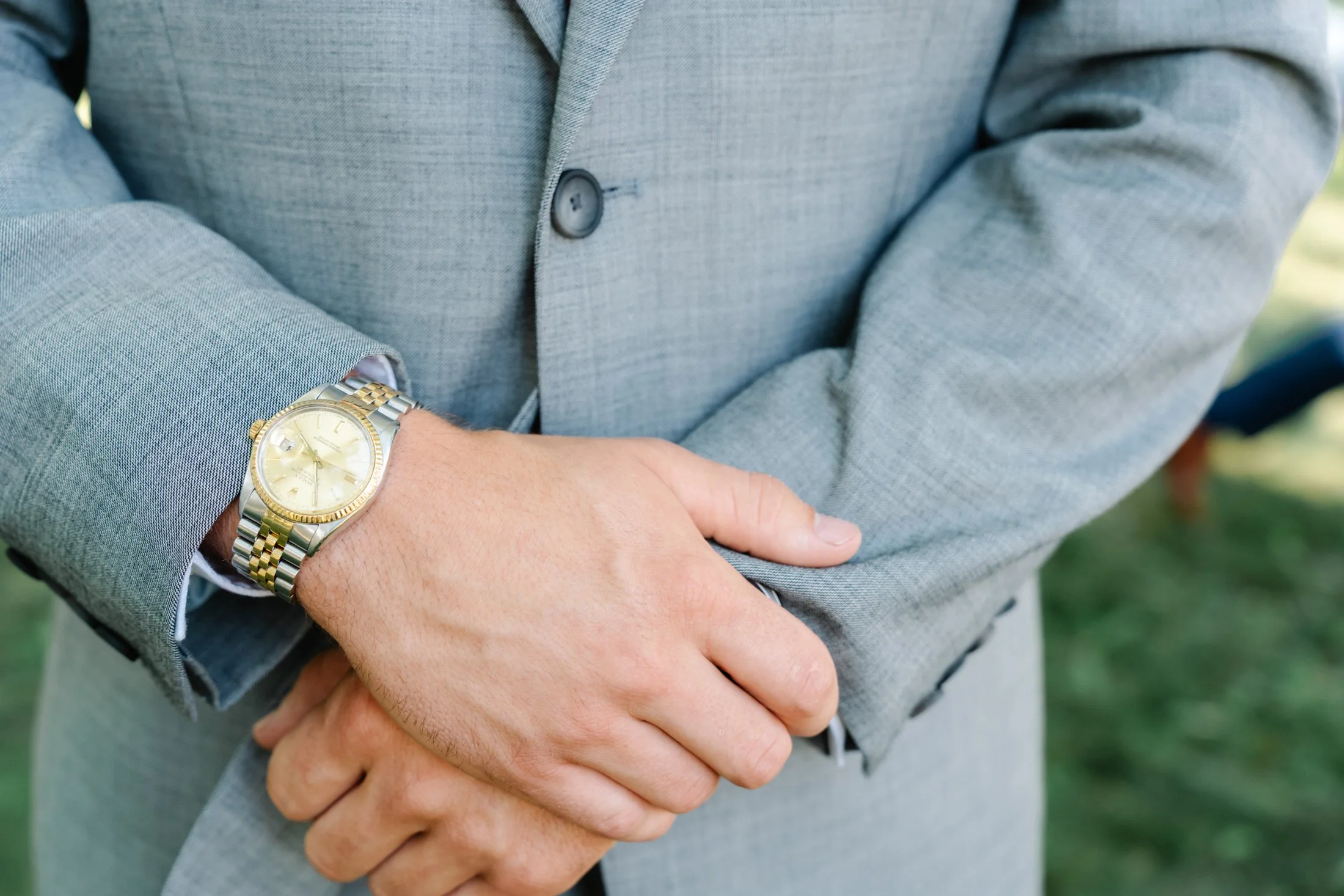Close-up of a person wearing a gray suit with a gold and silver wristwatch, standing outdoors with hands clasped.