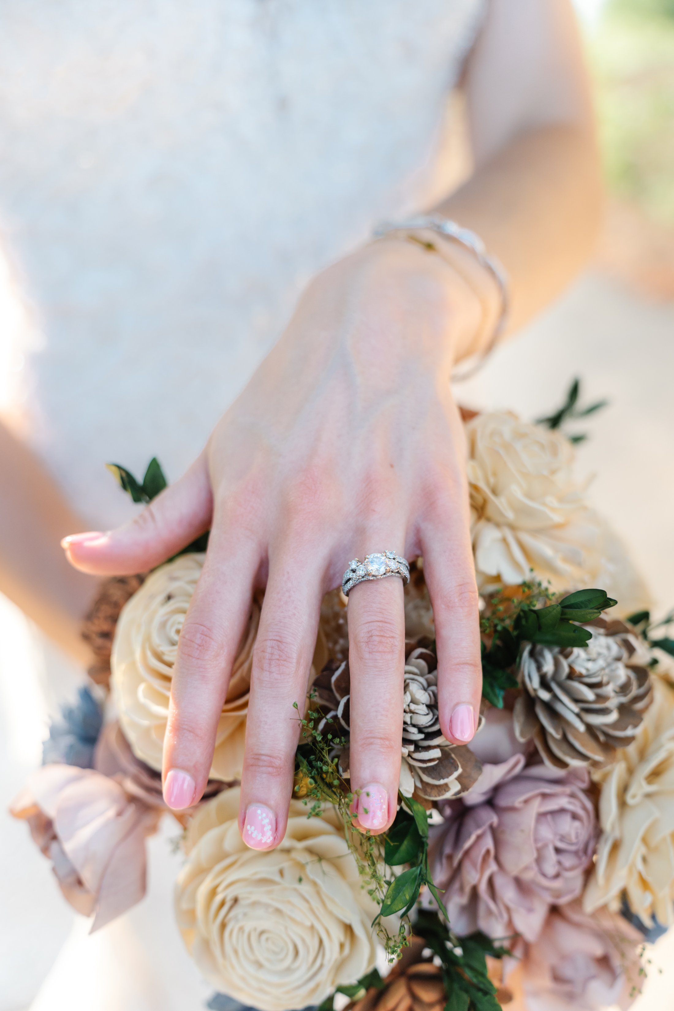 Close-up of a woman's left hand displaying a diamond engagement ring and wedding band, resting on a bouquet of roses and pinecones.