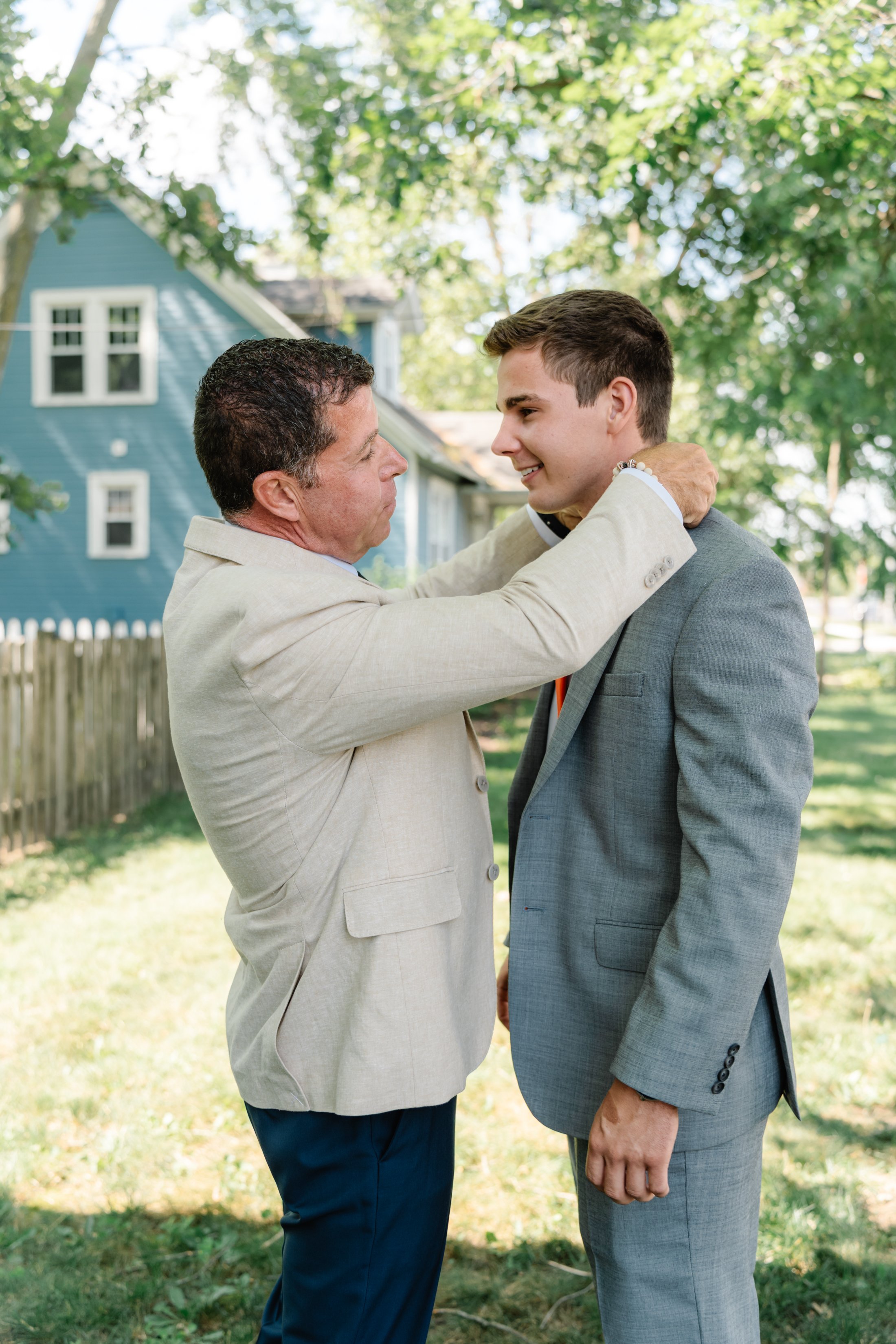 A man in a beige suit is holding a younger man in a gray suit by the shoulders, both smiling affectionately during an outdoor event on a sunny day with green trees and a house in the background.